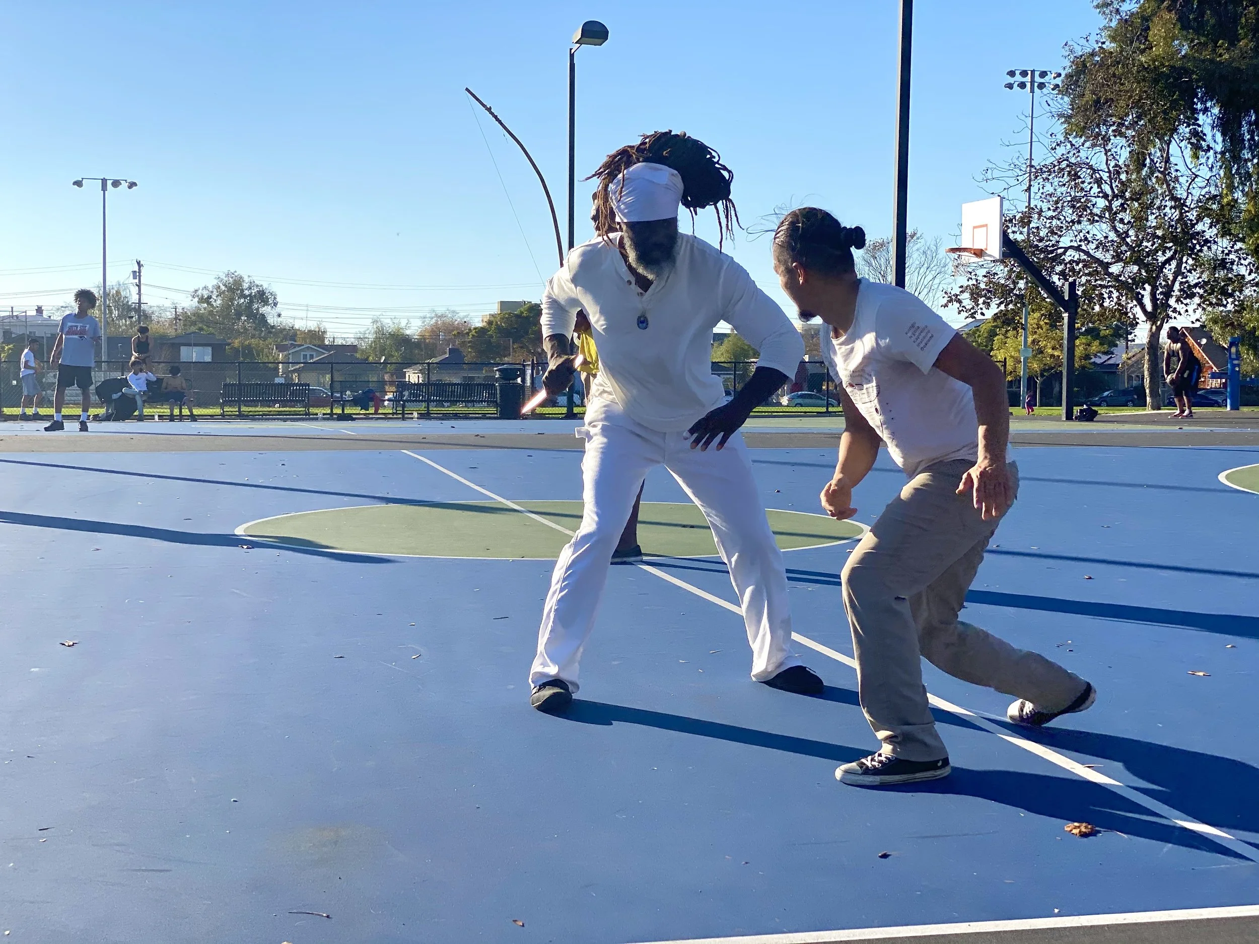 Capoeira de Angola instructor Nico Zuluaga and Treinel Mo of N'gola Preto Velho train Capoeira de Angola in Oakland, California outdoors on a sunny day. 