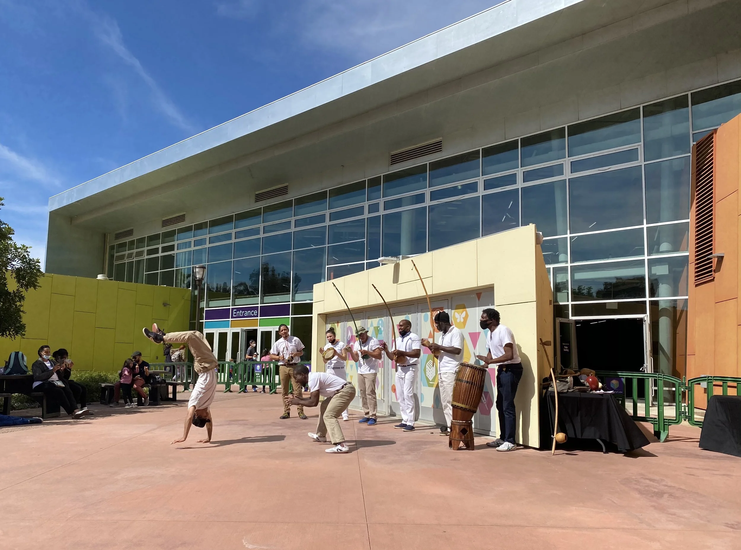 A group of performers performing capoeira, an Afro-Brazilian martial art, in front of a modern building with large glass windows. Some performers are mid-air flips, while others play drums and instruments, with an audience seated nearby.