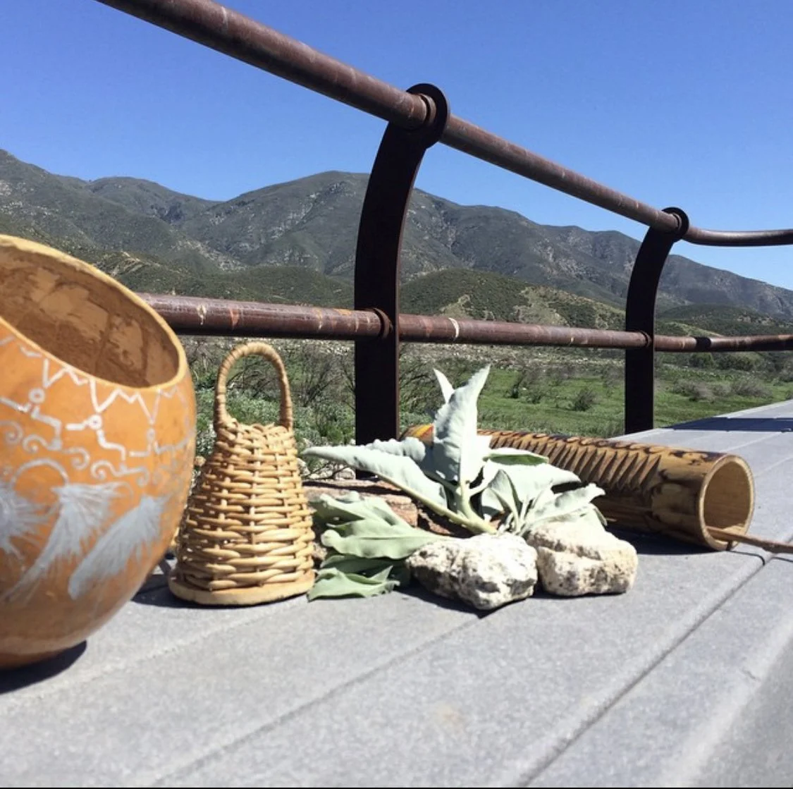 Outdoor scene with mountains in the background, a metal railing, and traditional capoeira instruments sitting next to ethically harvested White Sage in Southern California. 
