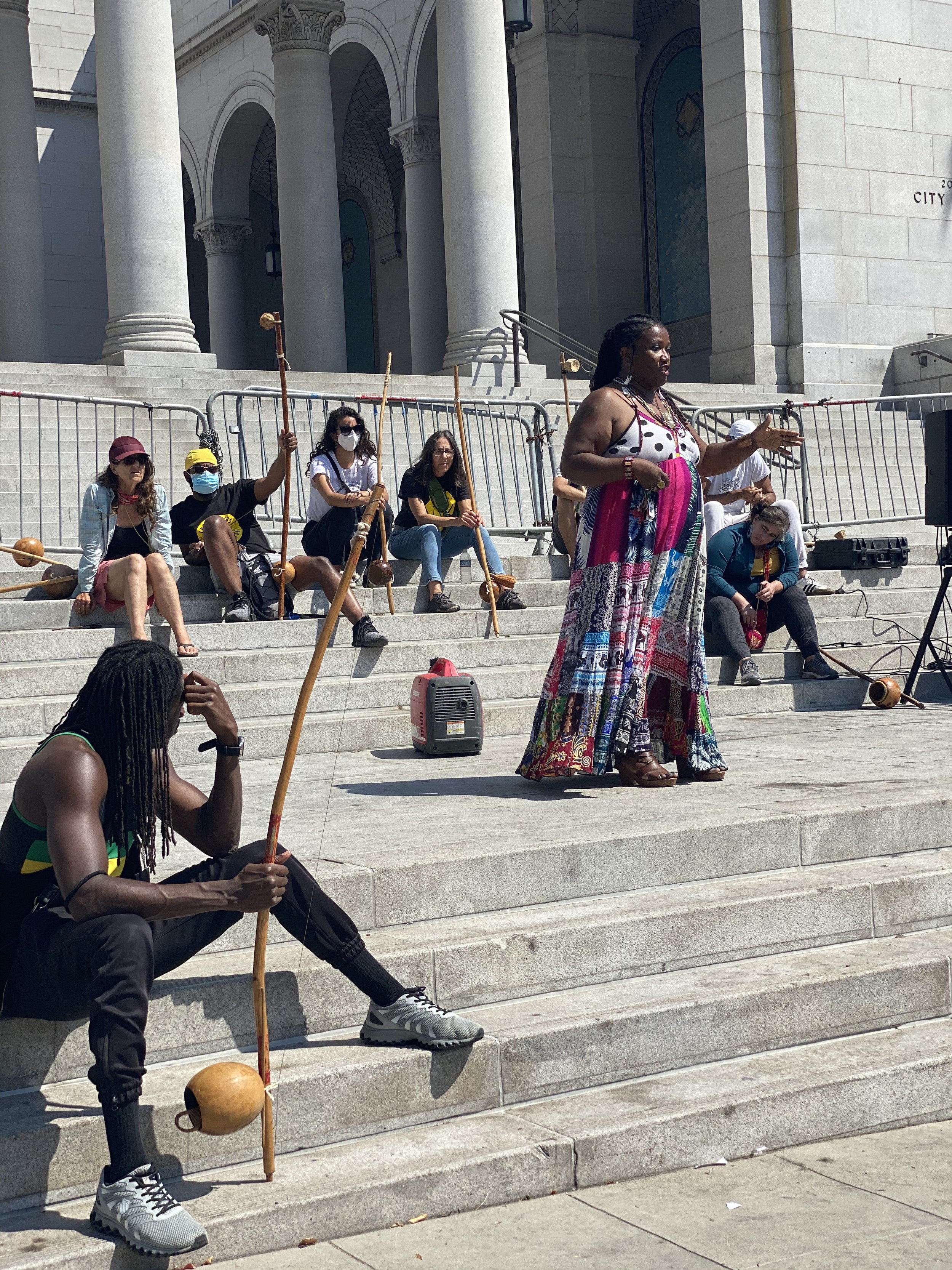 A woman in a colorful dress gives a speech to a group of people sitting on the steps of a historic building with large columns in downtown Los Angeles, California during a Capoeira de Angola presentation and community event.