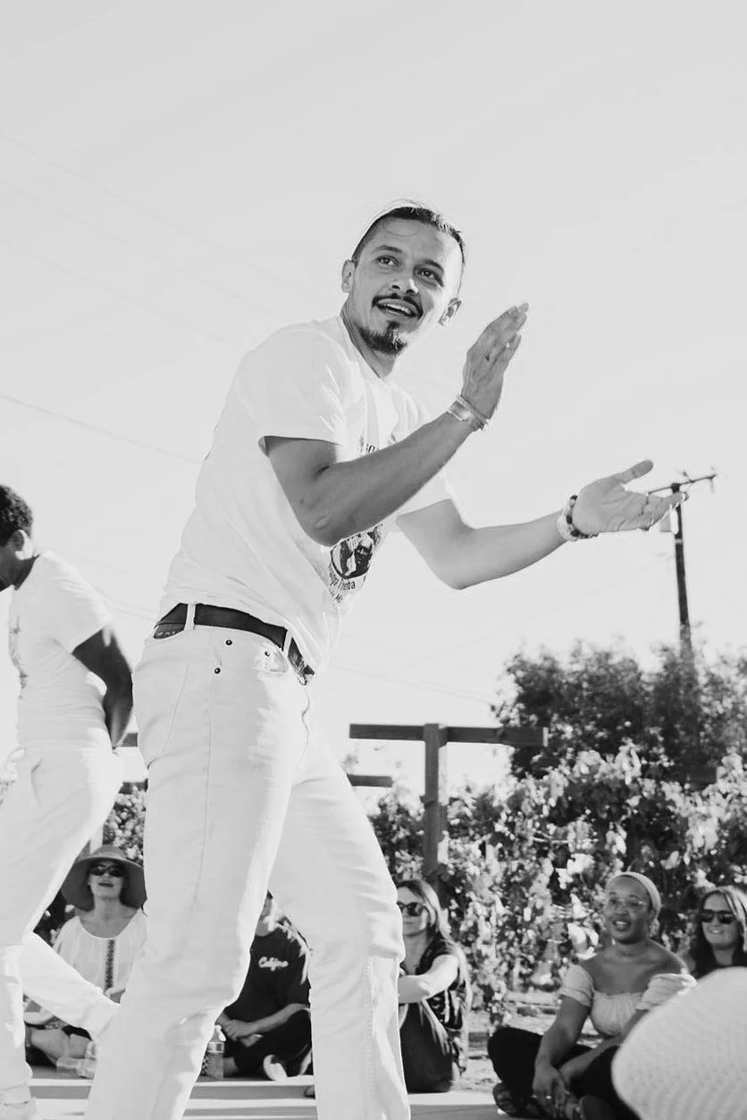 A man dancing outdoors with a group of smiling women seated on the ground, some wearing sunglasses and hats, while others smile and watch. The setting appears sunny with trees and a power line in the background.