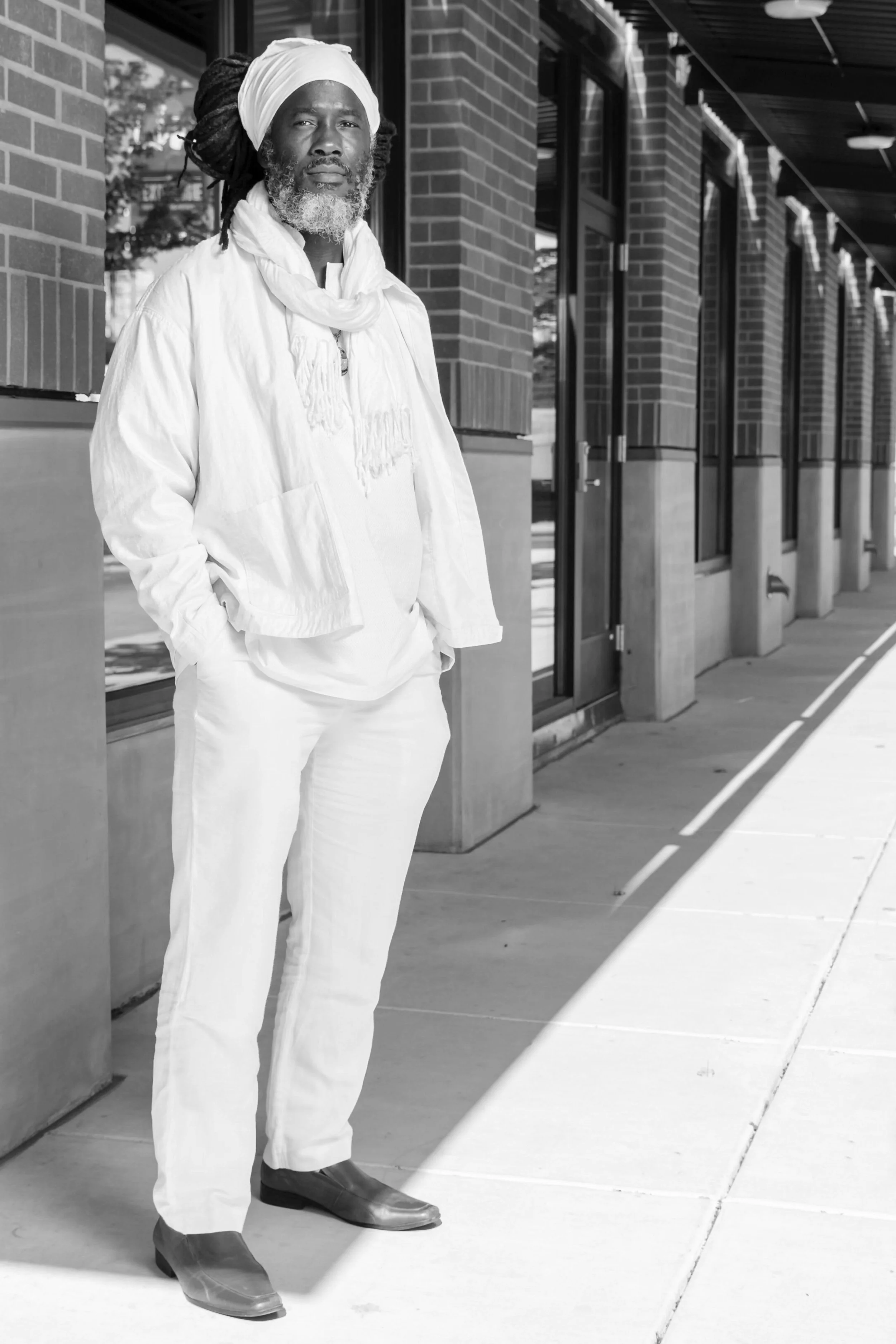 An older man with a beard and dreadlocks, wearing a white headwrap, scarf, jacket, and pants, stands on a sidewalk beside a brick building with large windows. He has his hands in his pockets and looks into the distance.