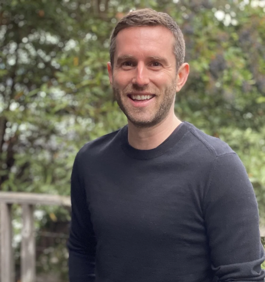 A smiling man with short brown hair and a beard, wearing a dark long-sleeve shirt, standing outdoors with trees in the background.