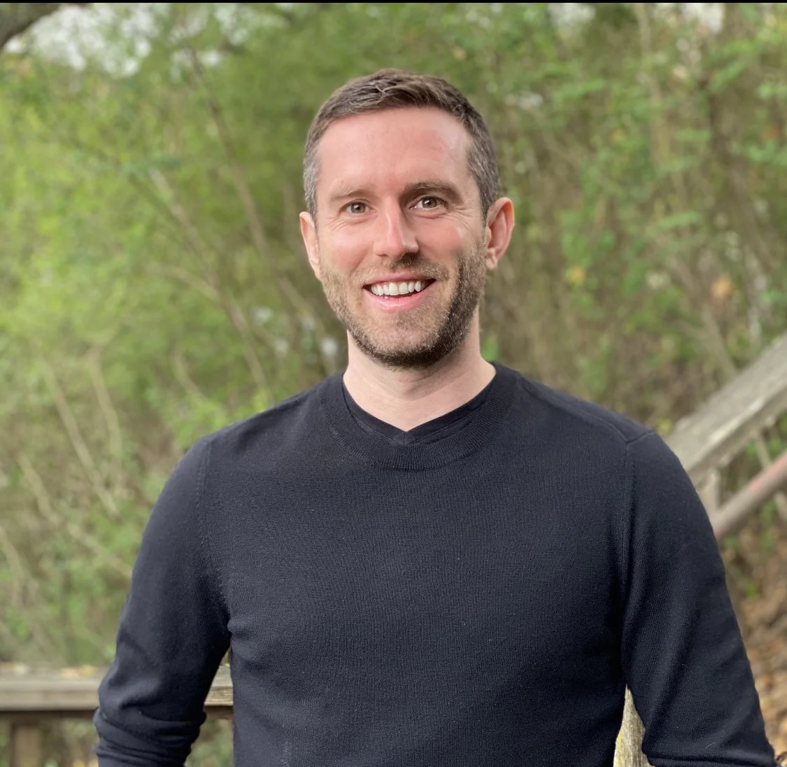 A smiling man with short brown hair and a beard, wearing a black sweater, outdoors with green trees in the background.