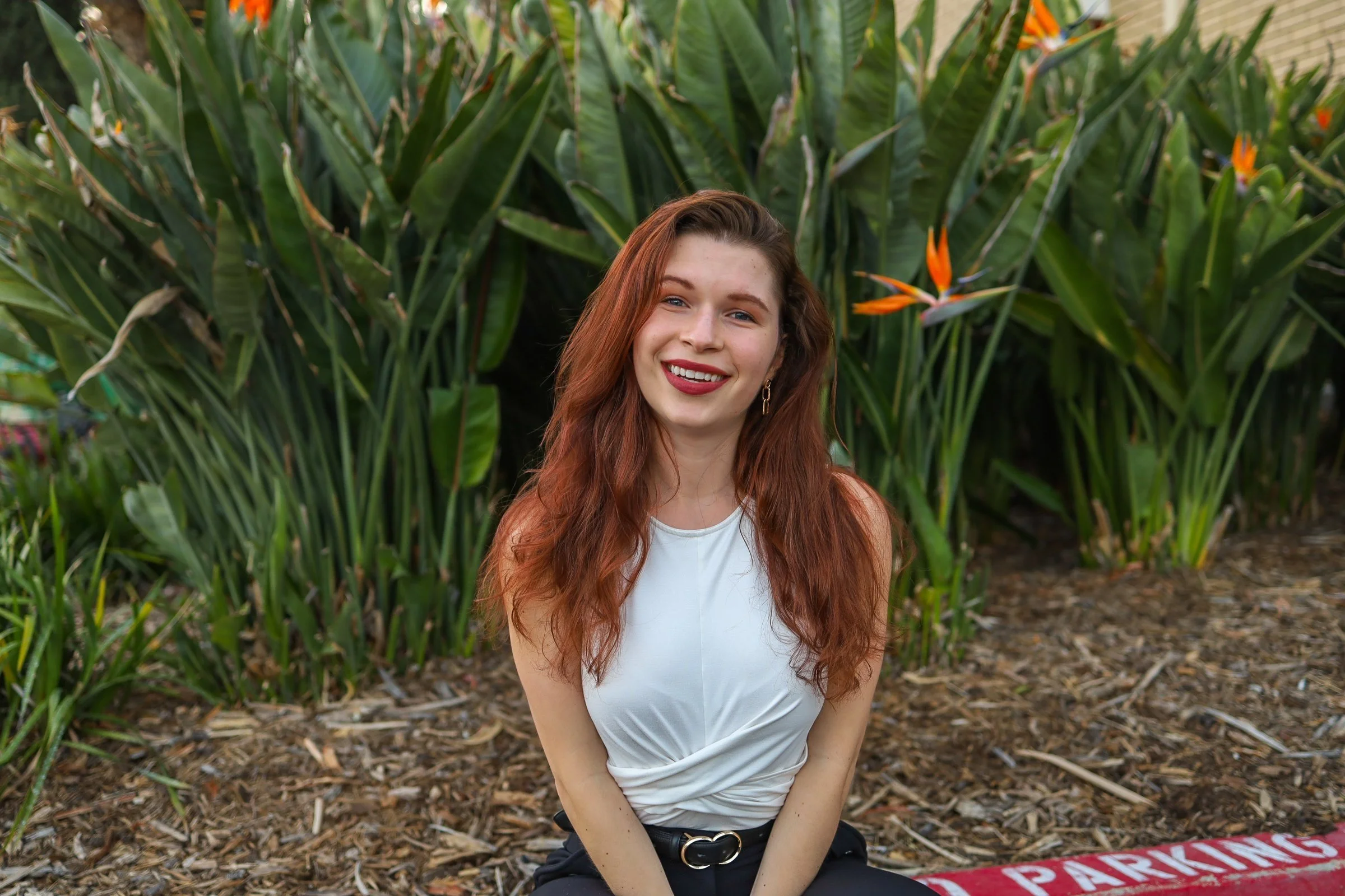 A young woman with long red hair and a white sleeveless top smiling while sitting in front of green tropical plants with orange flowers.