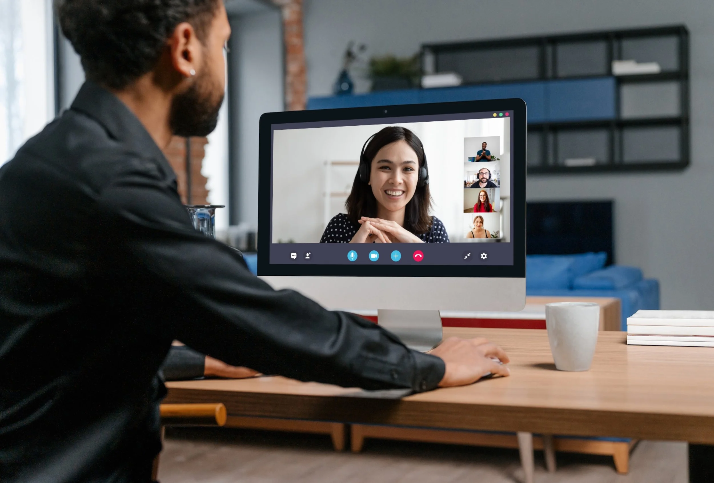 A person is sitting at a desk, participating in a video call on a desktop computer, with a woman smiling on the screen and four other participants in smaller video thumbnails.