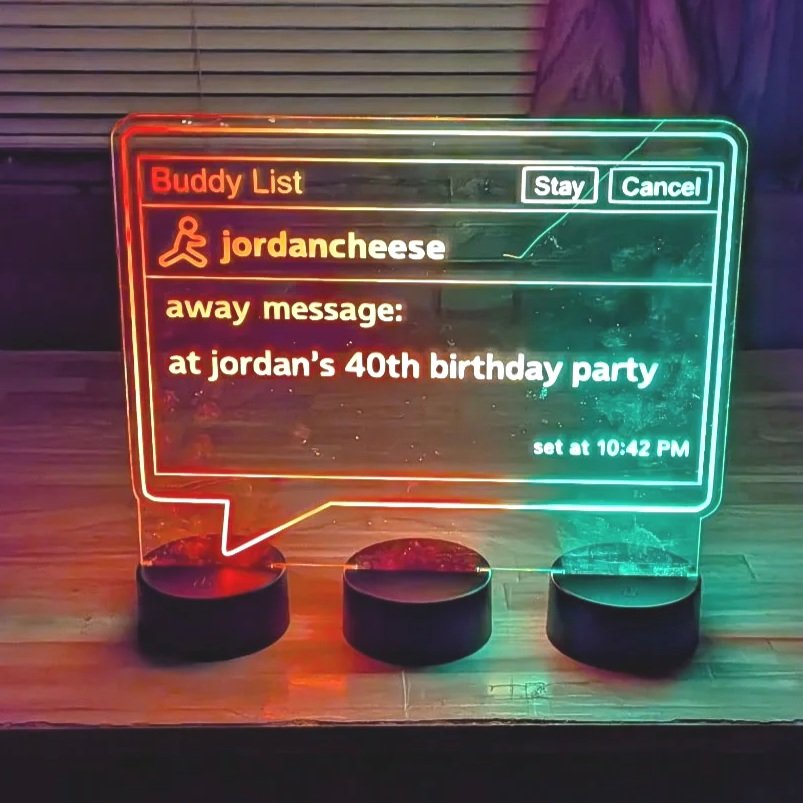 Colorful neon LED display with a digital message about a buddy list. The message states someone's 'away message' at a birthday party, with options to stay or cancel. The display is placed on a wooden surface.