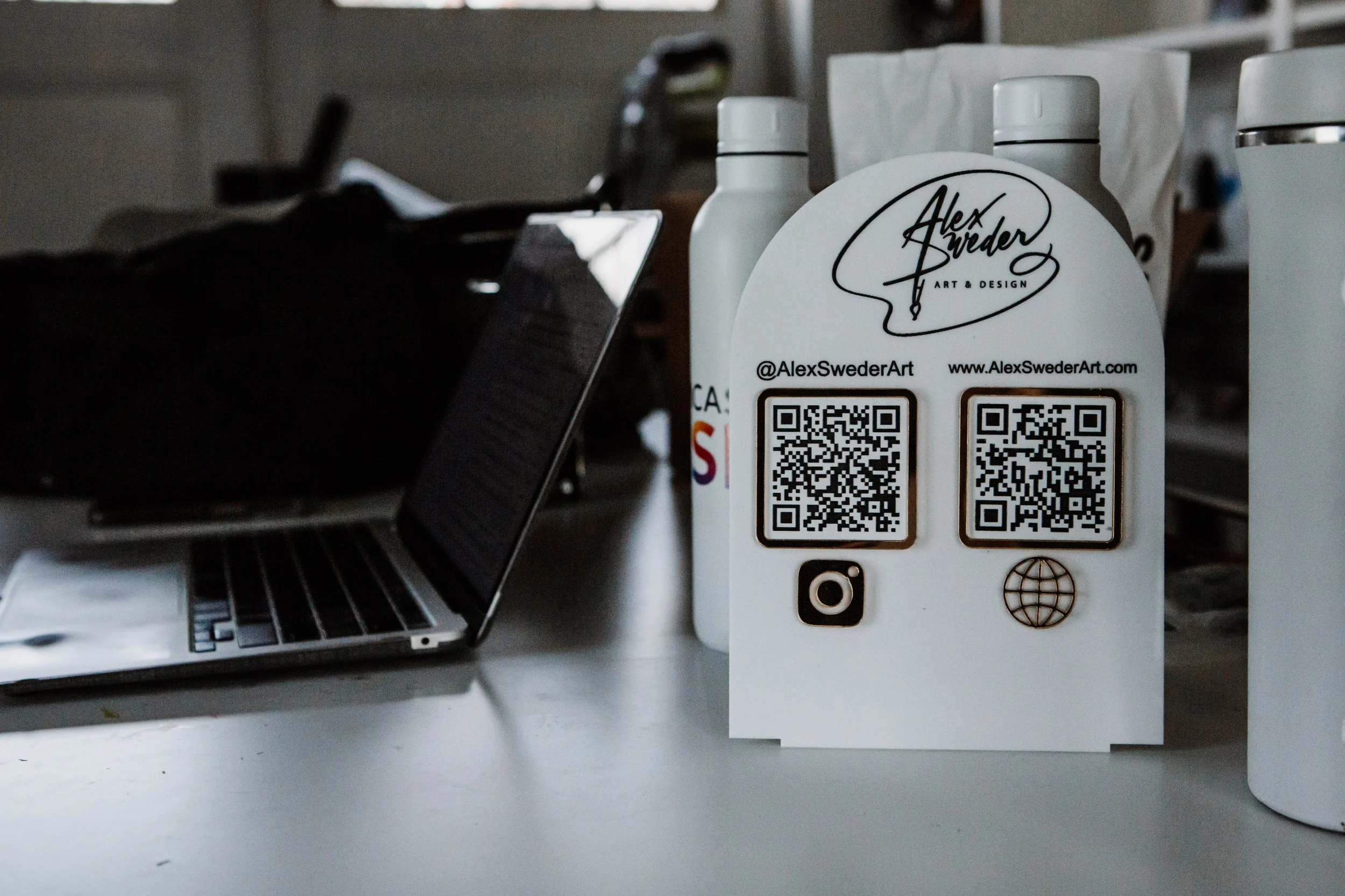 Business card with signature logo, social media icons, and QR codes on a white surface near a laptop and white bottles.