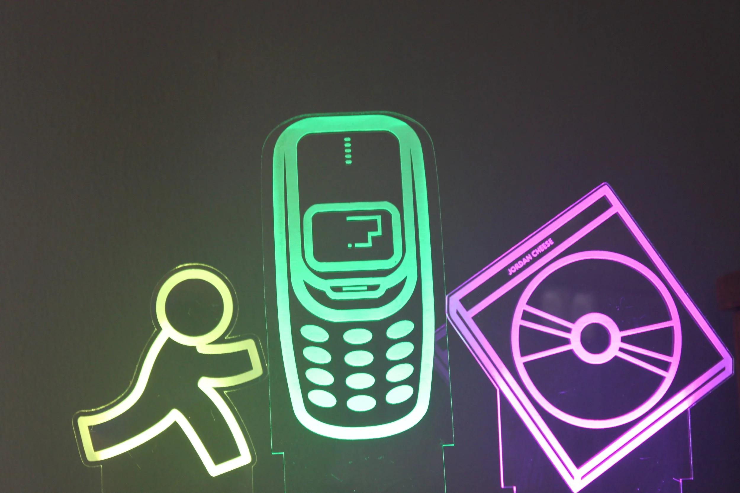Neon neon signs of a person, a mobile phone with a question mark on the screen, and a CD or DVD disc with the name Jordan Chee in pink.