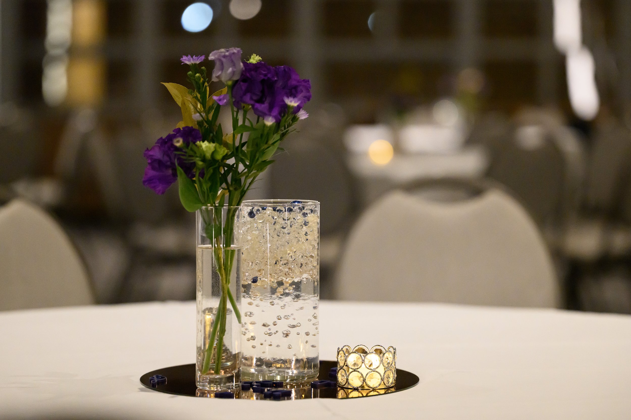 A table centerpiece with a tall glass vase holding purple and white flowers, a smaller glass container with bubbles, and a decorative candle holder with tealights, on a black round tray, in a dimly lit event setting.