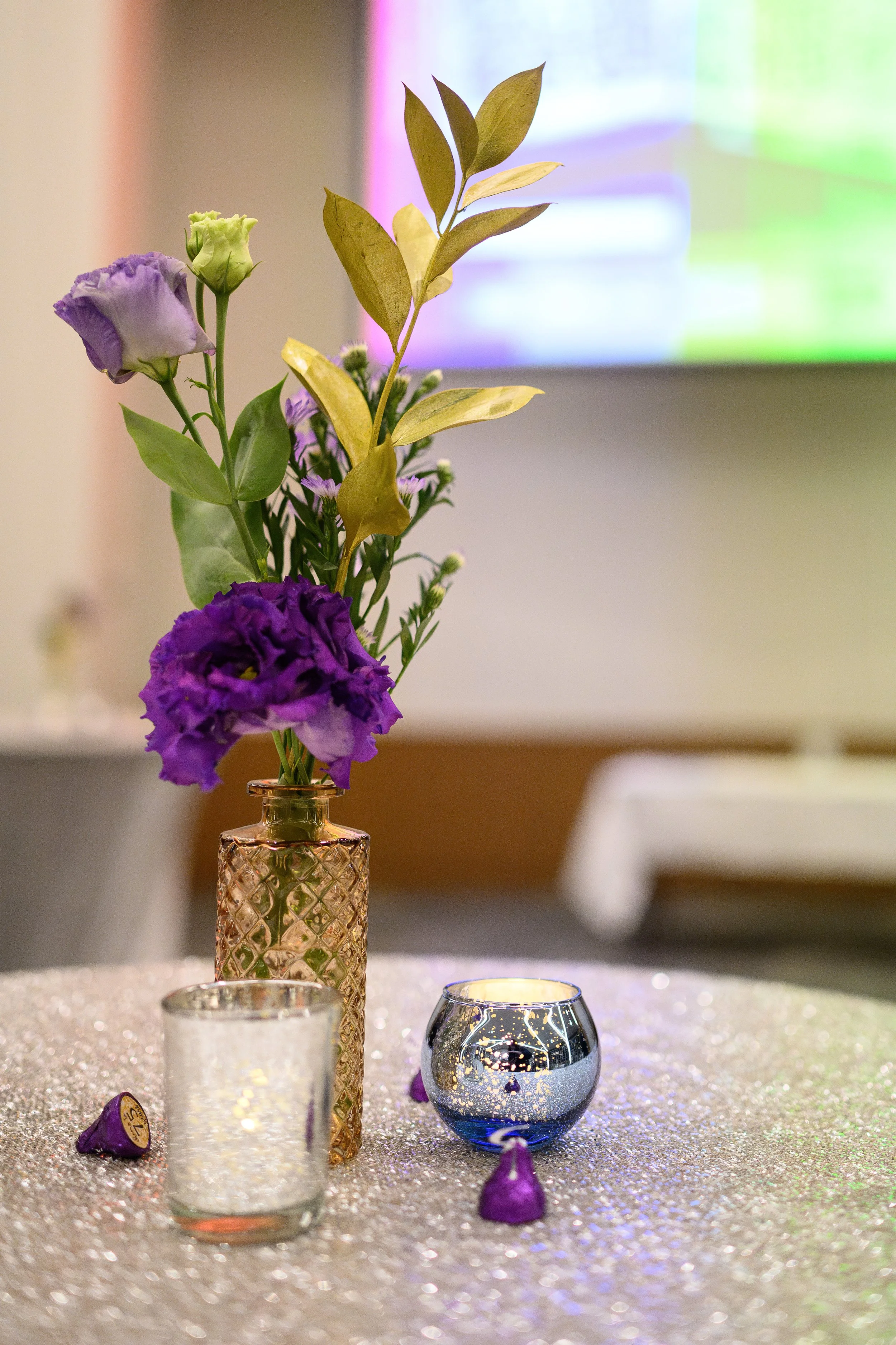 A small bouquet of purple and white flowers in a pink textured glass vase, with a purple tealight holder and two small purple candies on a glittery tablecloth.