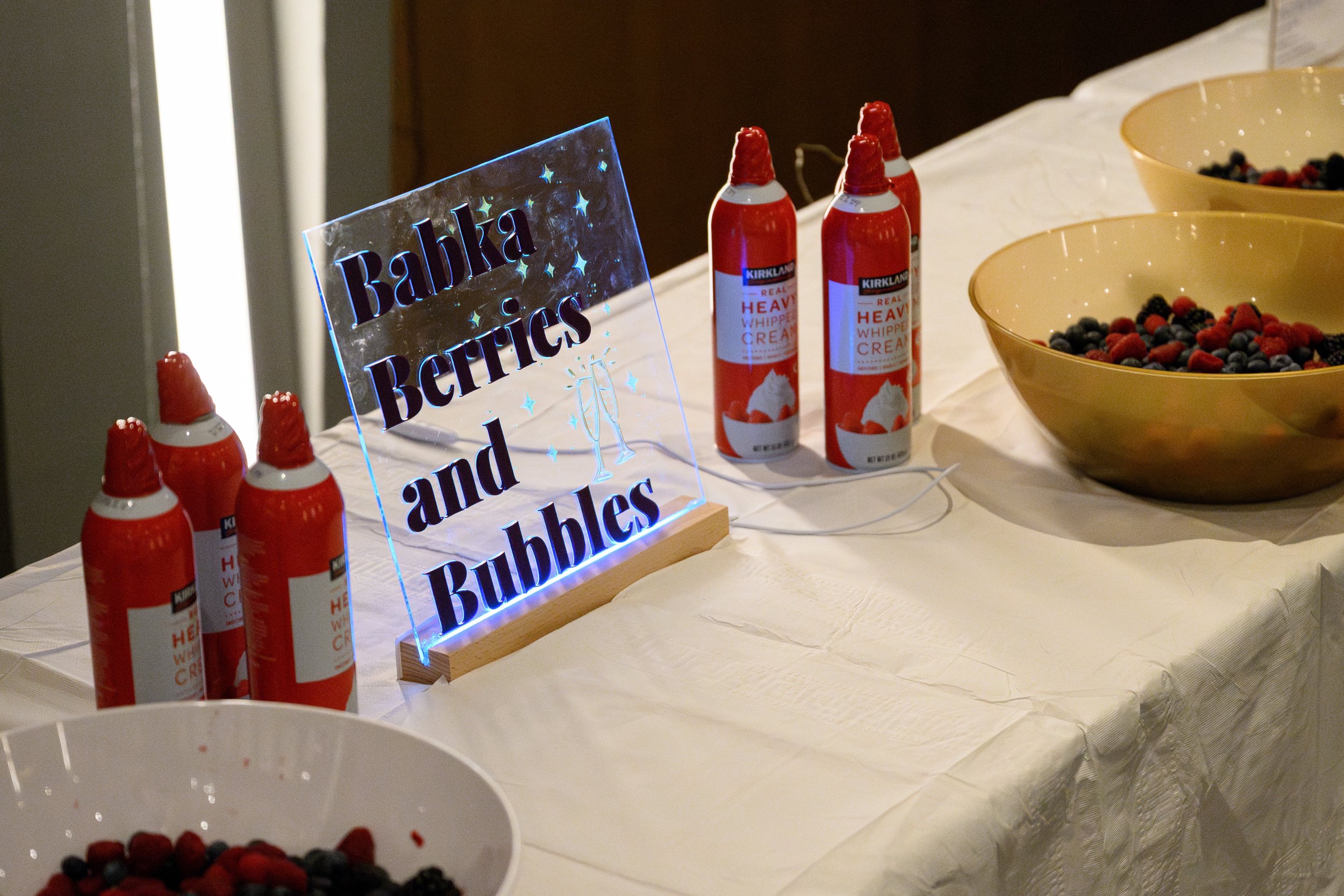 Table with bowls of mixed berries, cans of whipped cream, and a lit sign that reads 'Babka Berries and Bubbles'