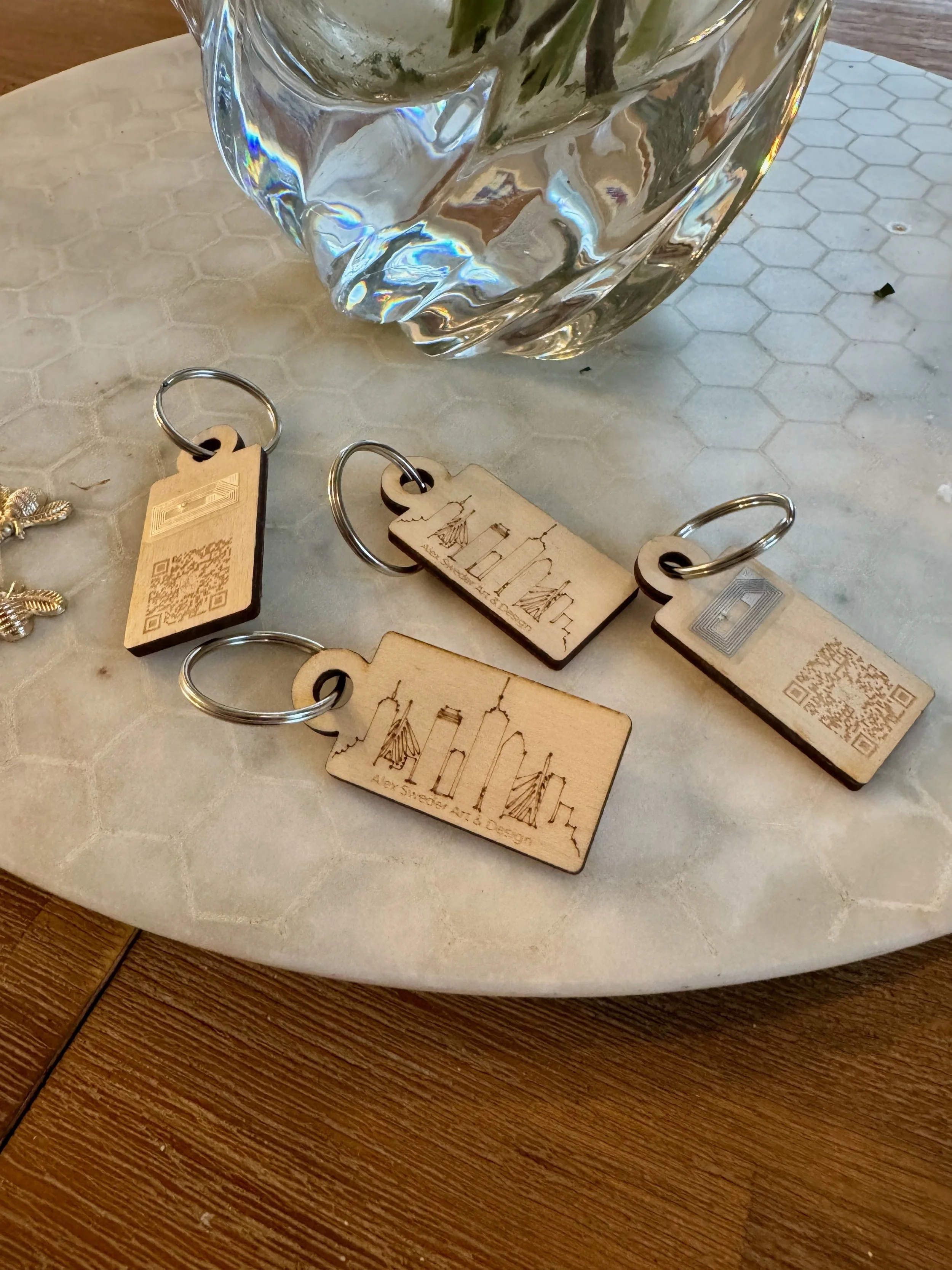 Three wooden keychains with New York City skyline engravings, placed on a table with a mosaic pattern and a glass vase in the background.