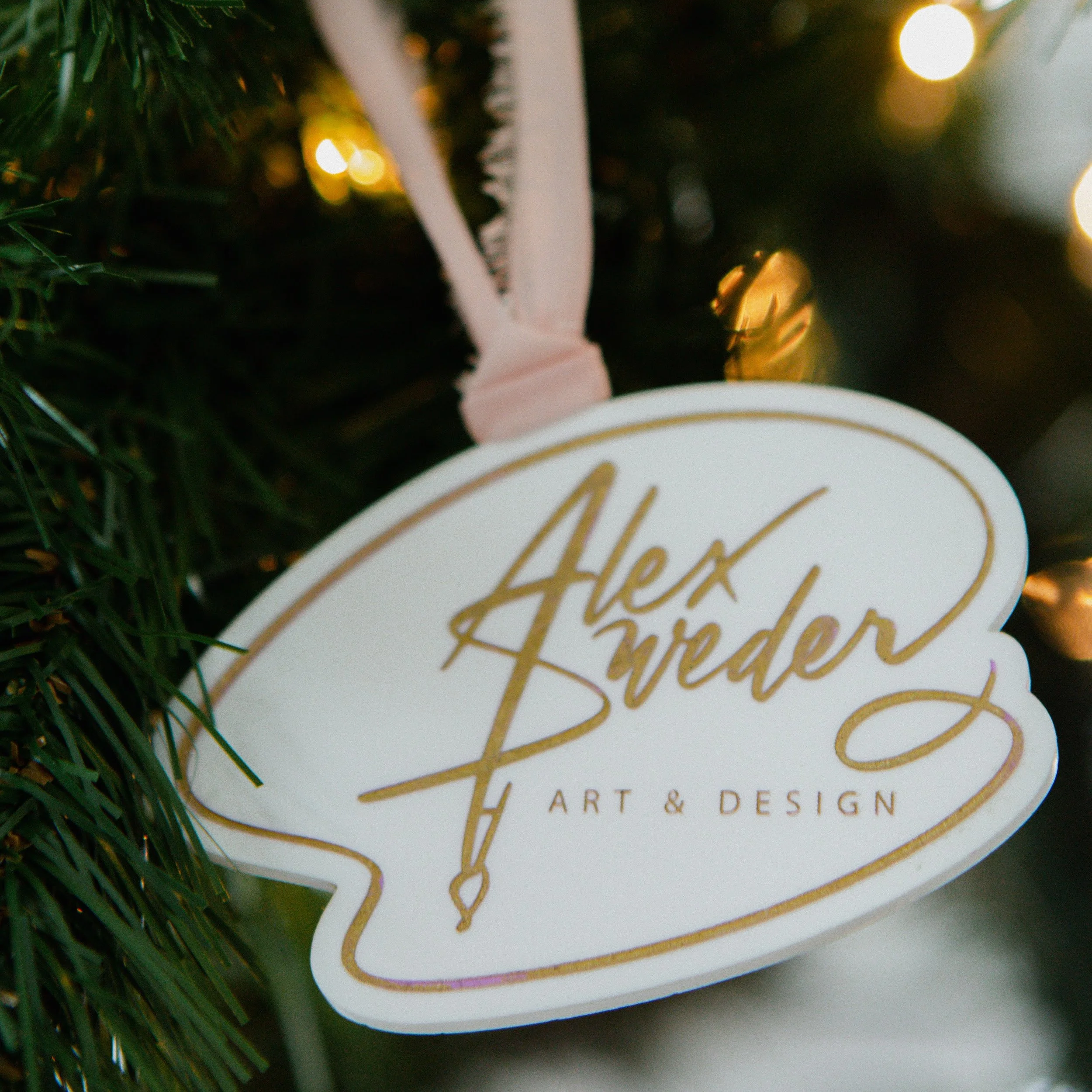 A Christmas tree ornament with the name 'Alex Sweder' and 'Art & Design' written in gold on a white background, hanging from a pink ribbon among Christmas tree branches and lights.