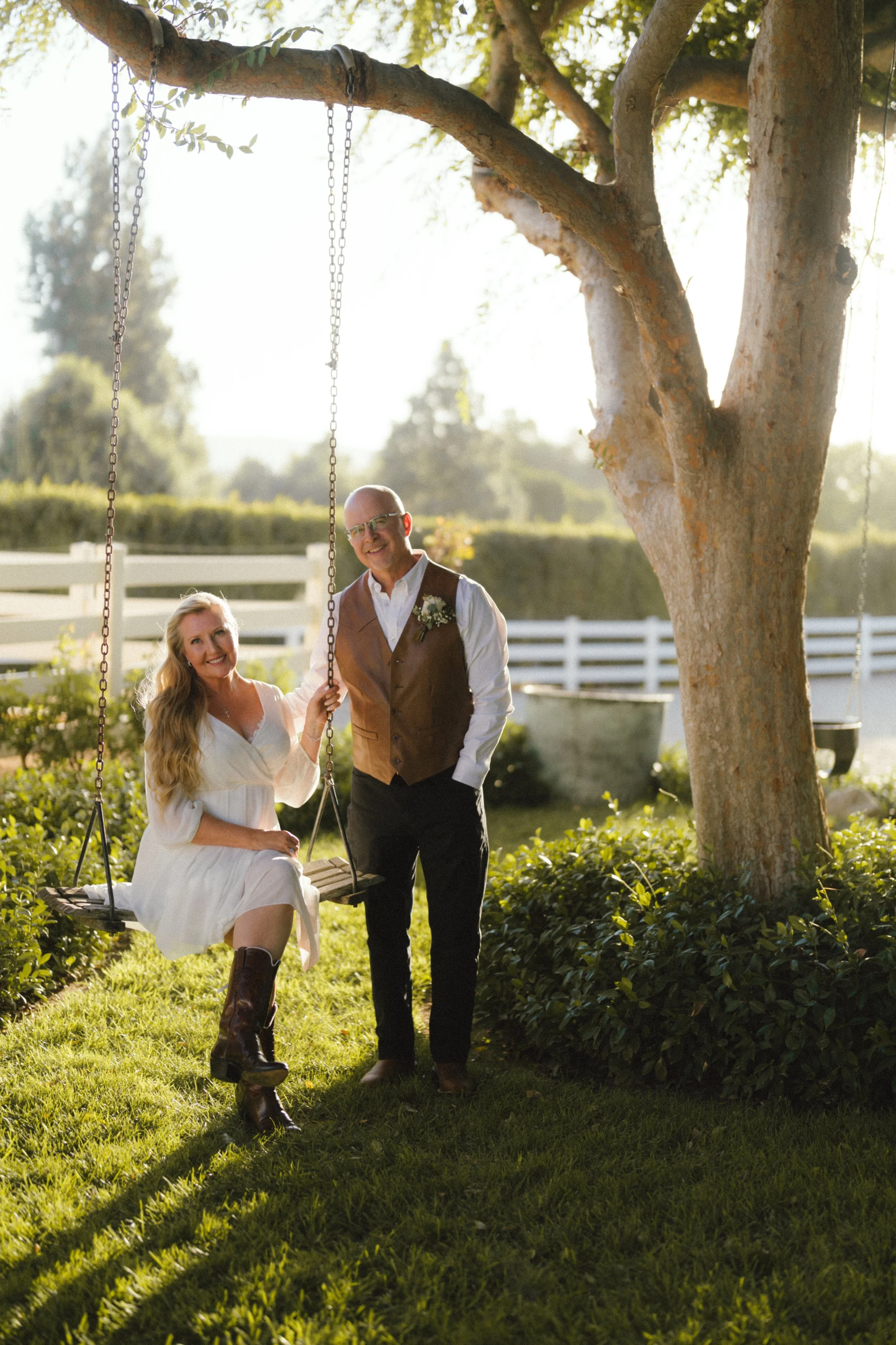 A woman sitting on a wooden swing hanging from a tree, with a man standing beside her, both smiling outdoors in a garden with a white fence and lush greenery.