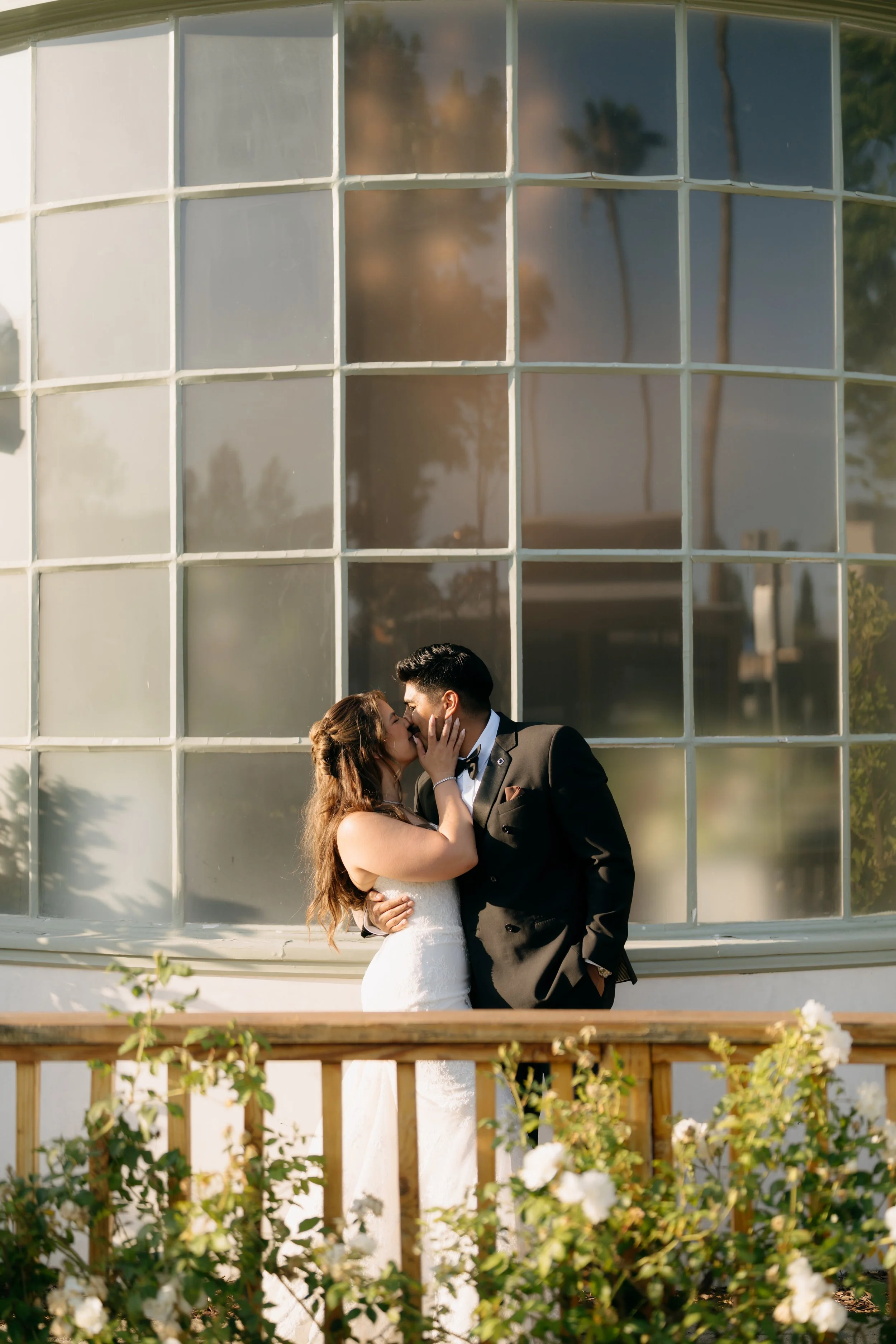 A wedding couple sharing a kiss outside a building with large grid-pane windows, surrounded by flowering bushes and greenery.