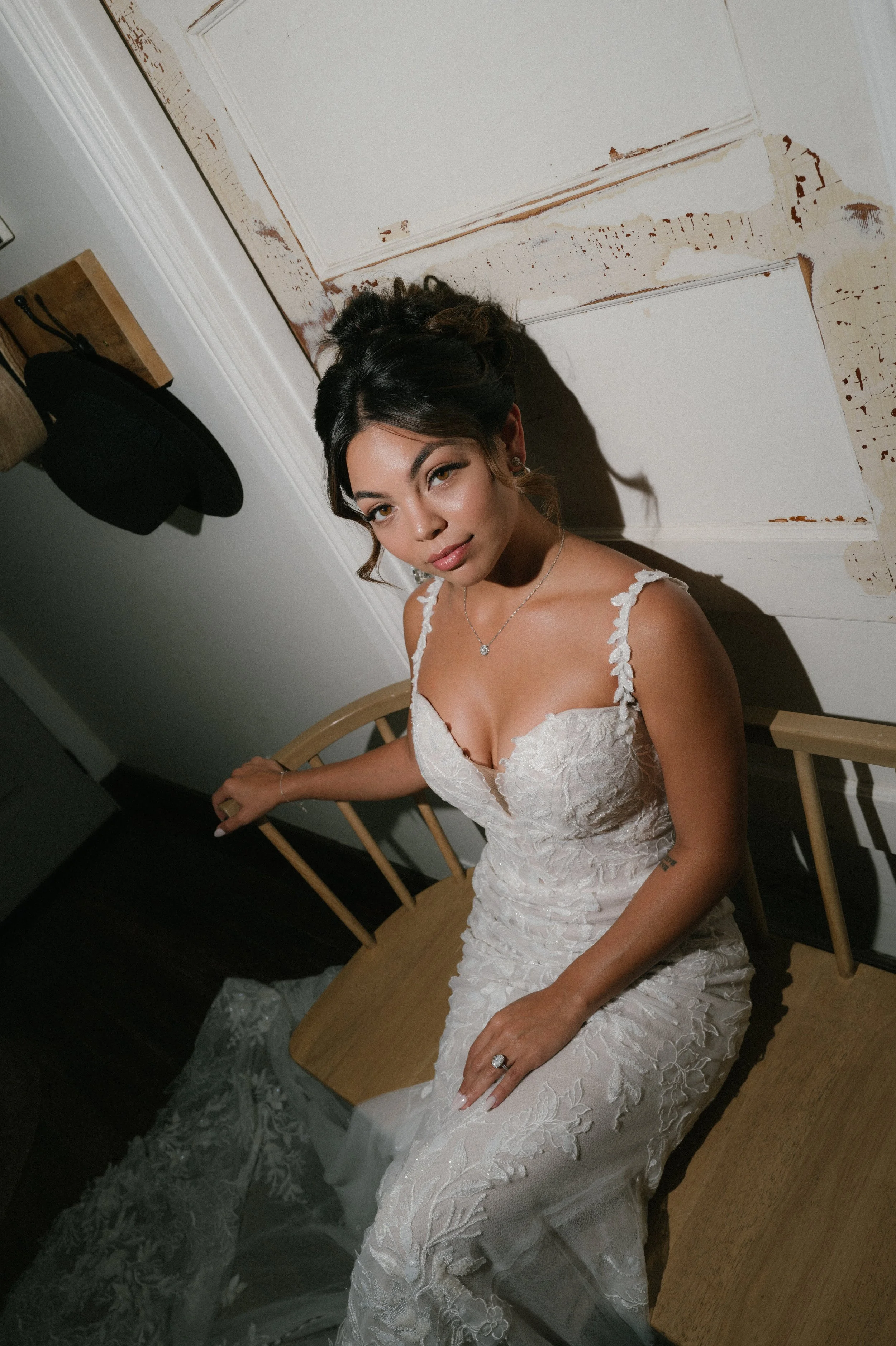 A woman in a white lace wedding dress sitting on a wooden chair, looking at the camera, with dark hair styled up, in a room with a white door and a hat hanging on the wall.