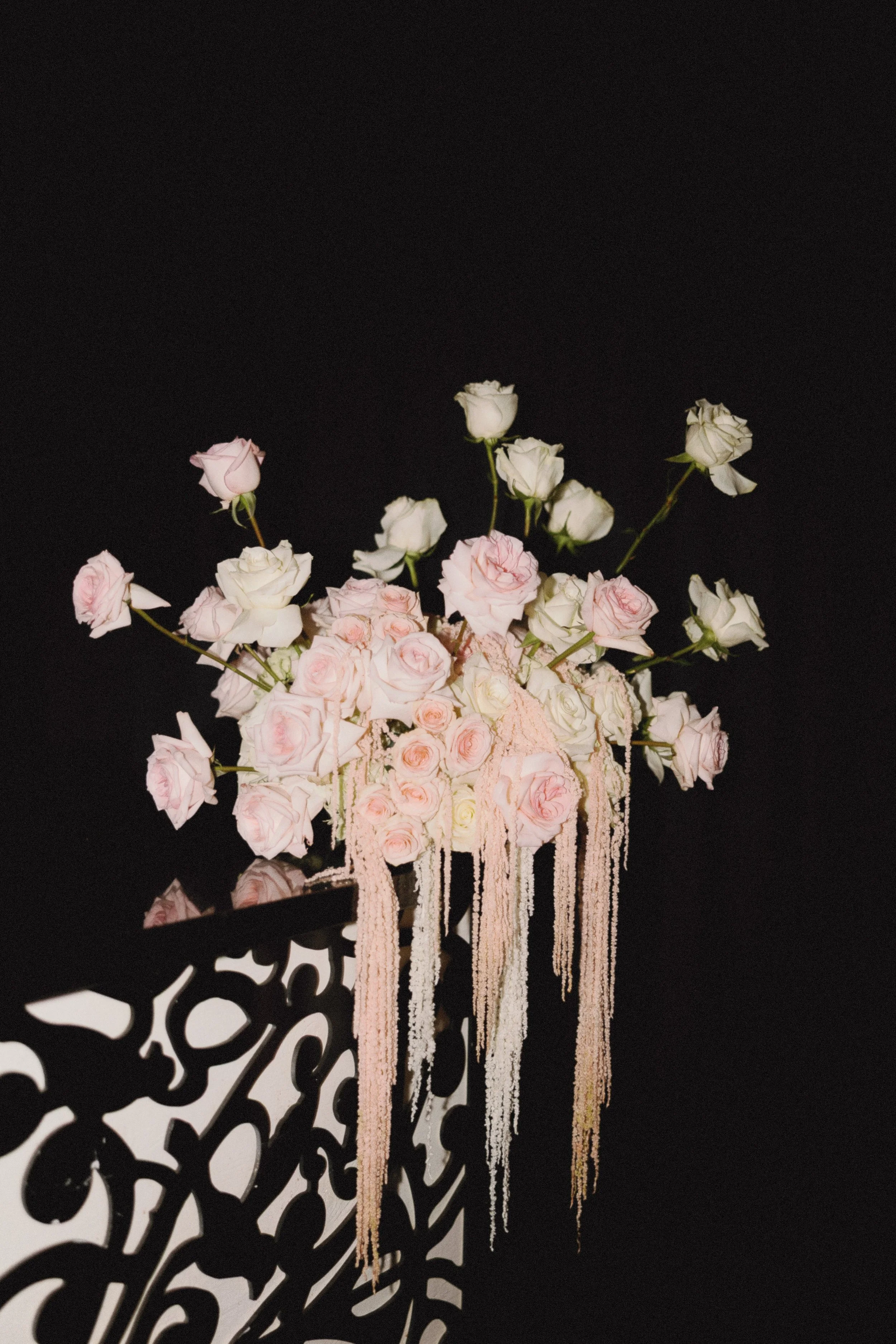 A black background with a floral arrangement of pink and white roses, decorated with long, hanging pink and white strands, placed on a black decorative stand.