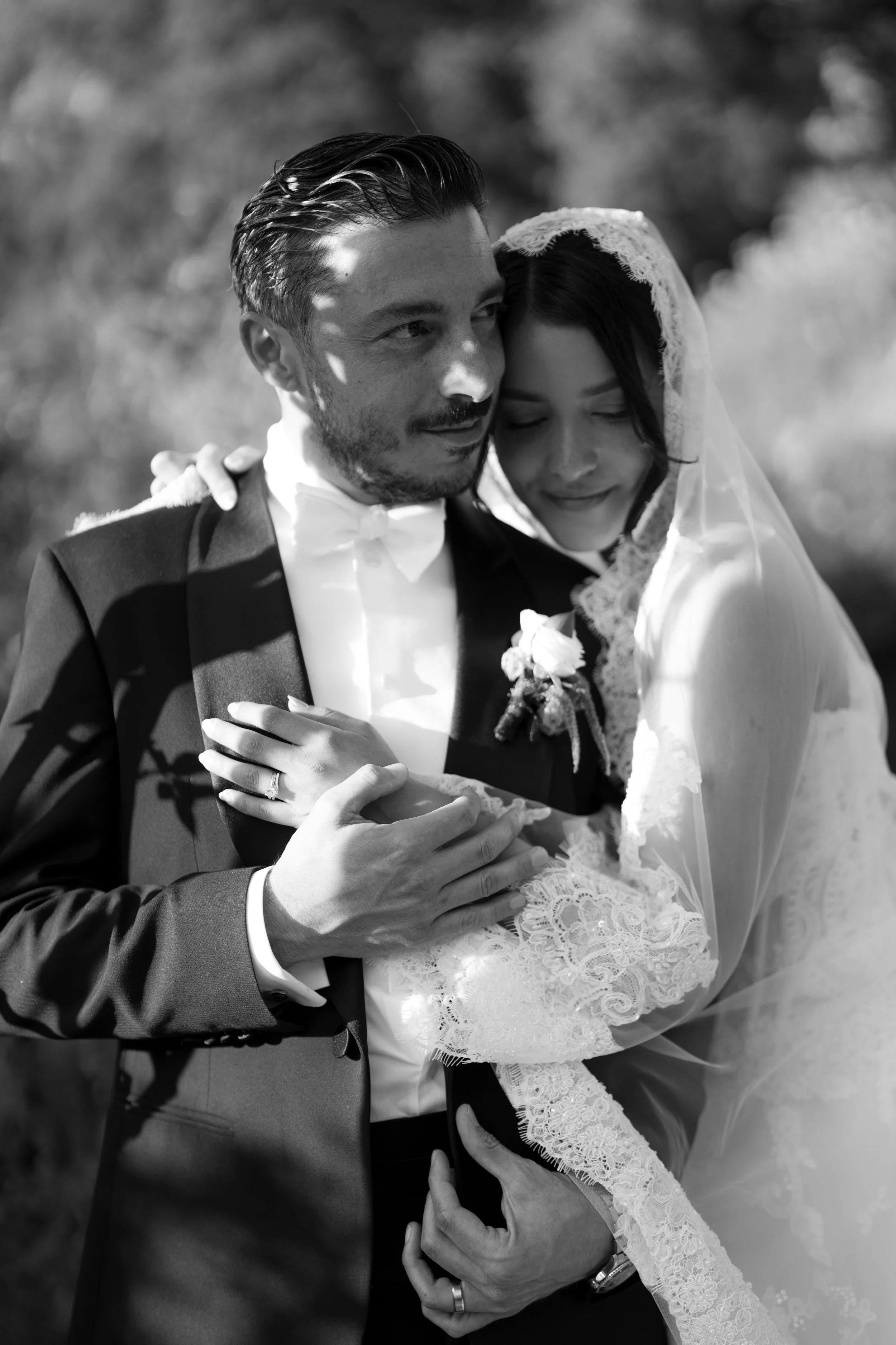 Black and white photo of a groom and bride embracing, with the groom in a tuxedo and the bride in a lace dress with a veil, both with their eyes closed.