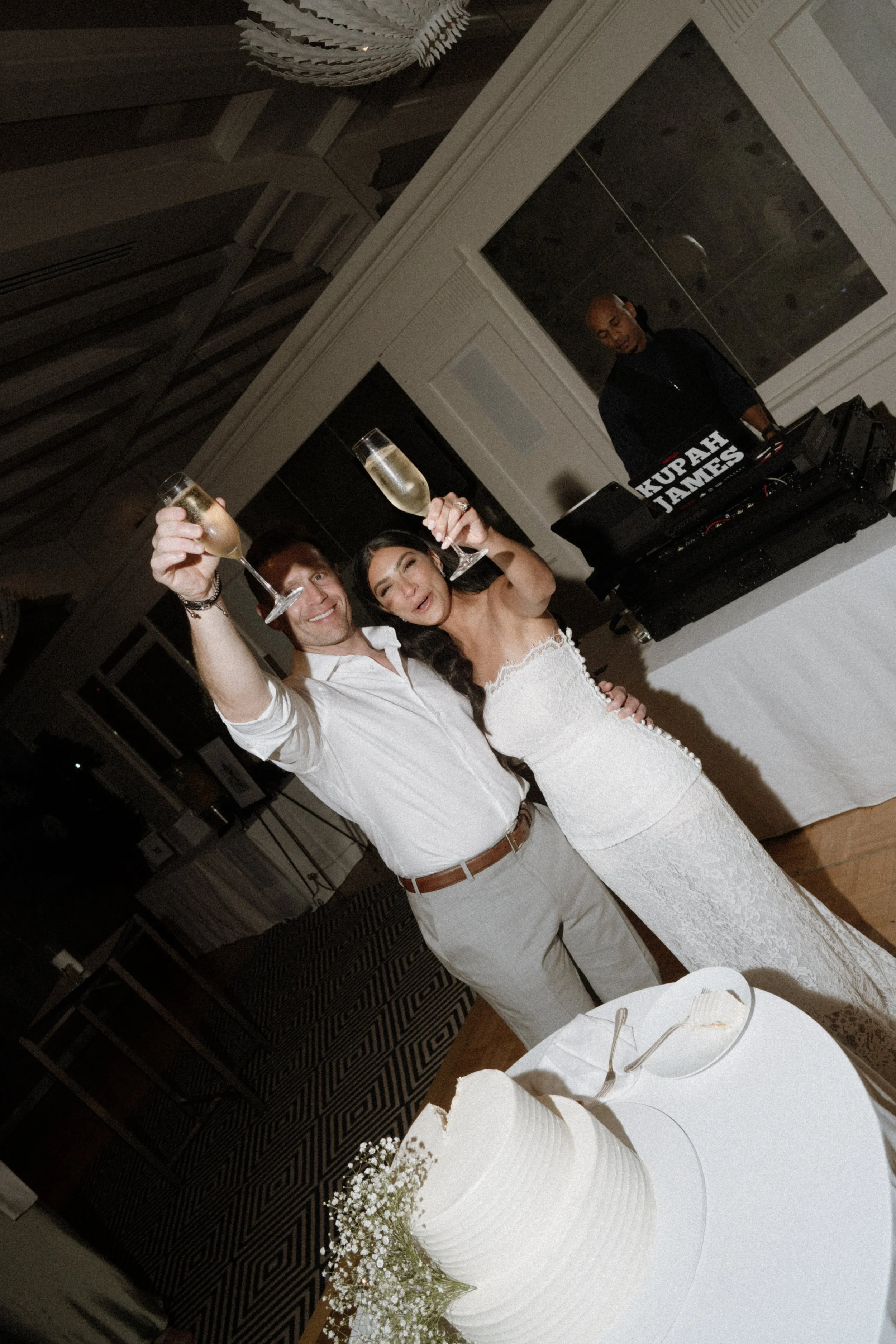 A man and woman at a celebration, holding champagne glasses and smiling. The woman is wearing a white dress, and the man is in a white shirt with sleeves rolled up. They are standing near a white cake decorated with baby's breath flowers. In the back