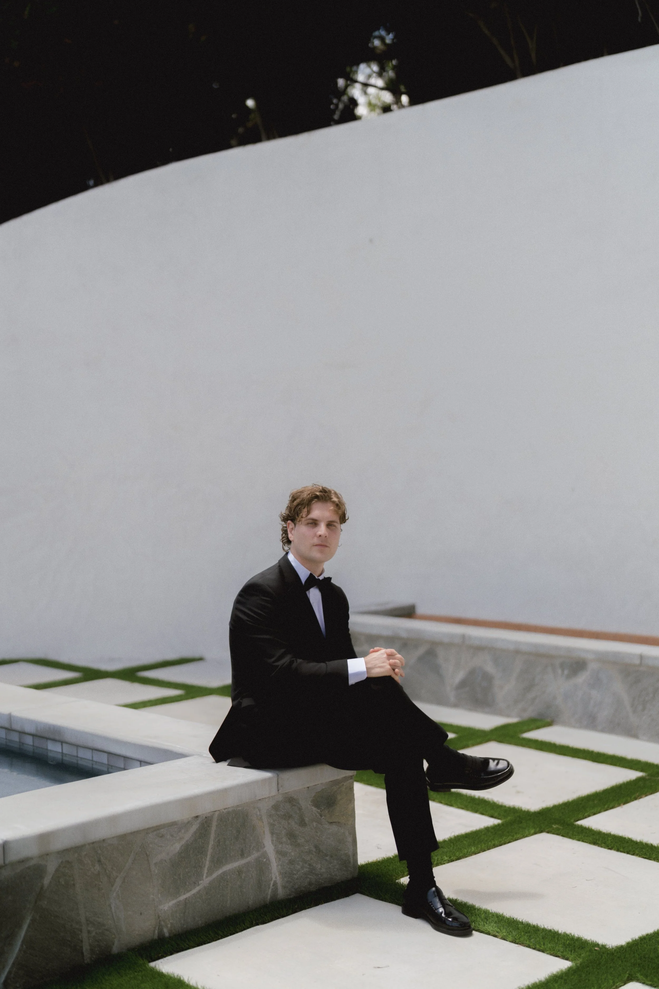 A man dressed in a black tuxedo, sitting on a stone bench outdoors, in front of a modern white building with a pool in the foreground and decorative grass patches.