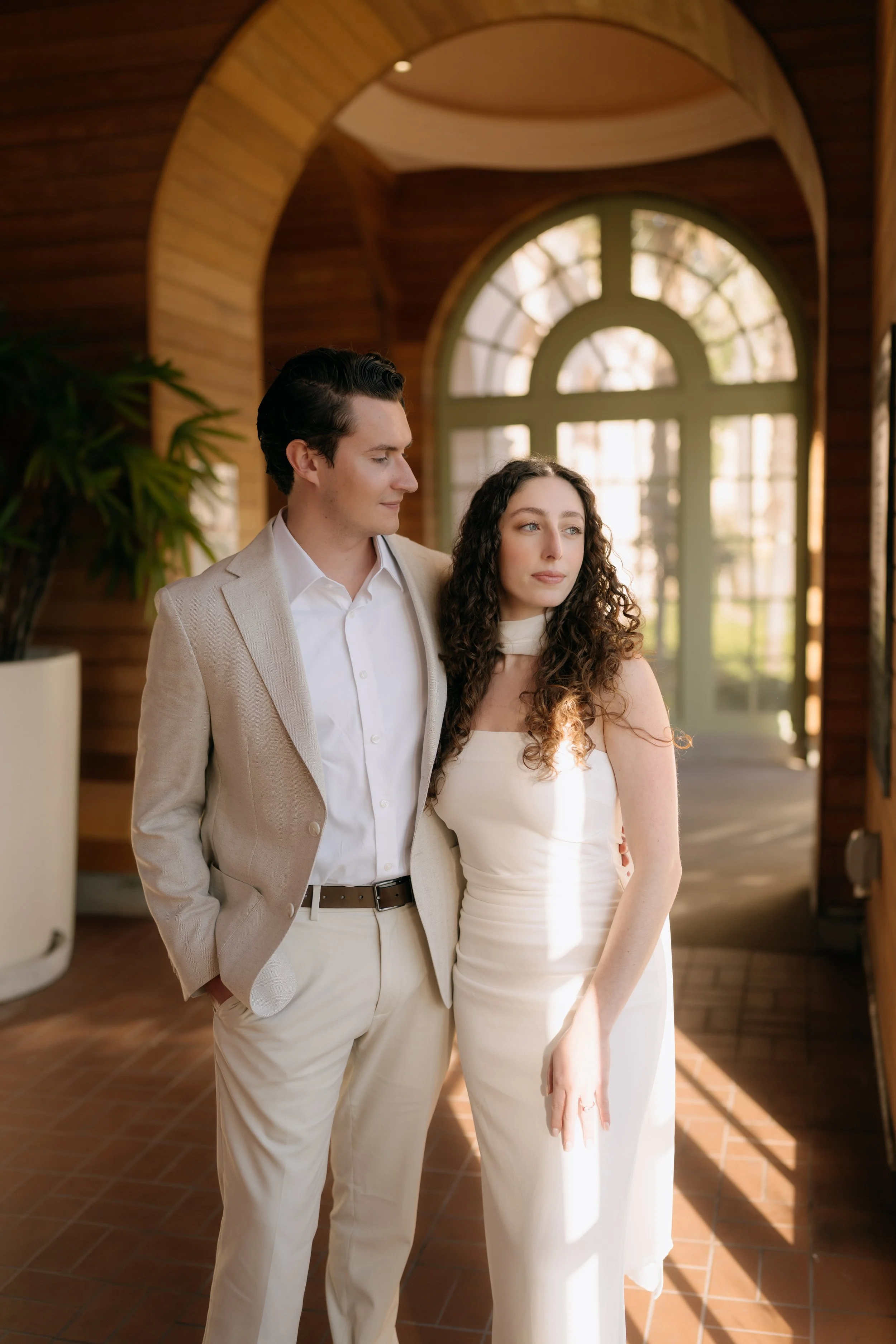 A young man and woman standing closely together inside a warmly lit, modern building with wooden walls and large arched windows, both dressed in elegant, light-colored formal attire.