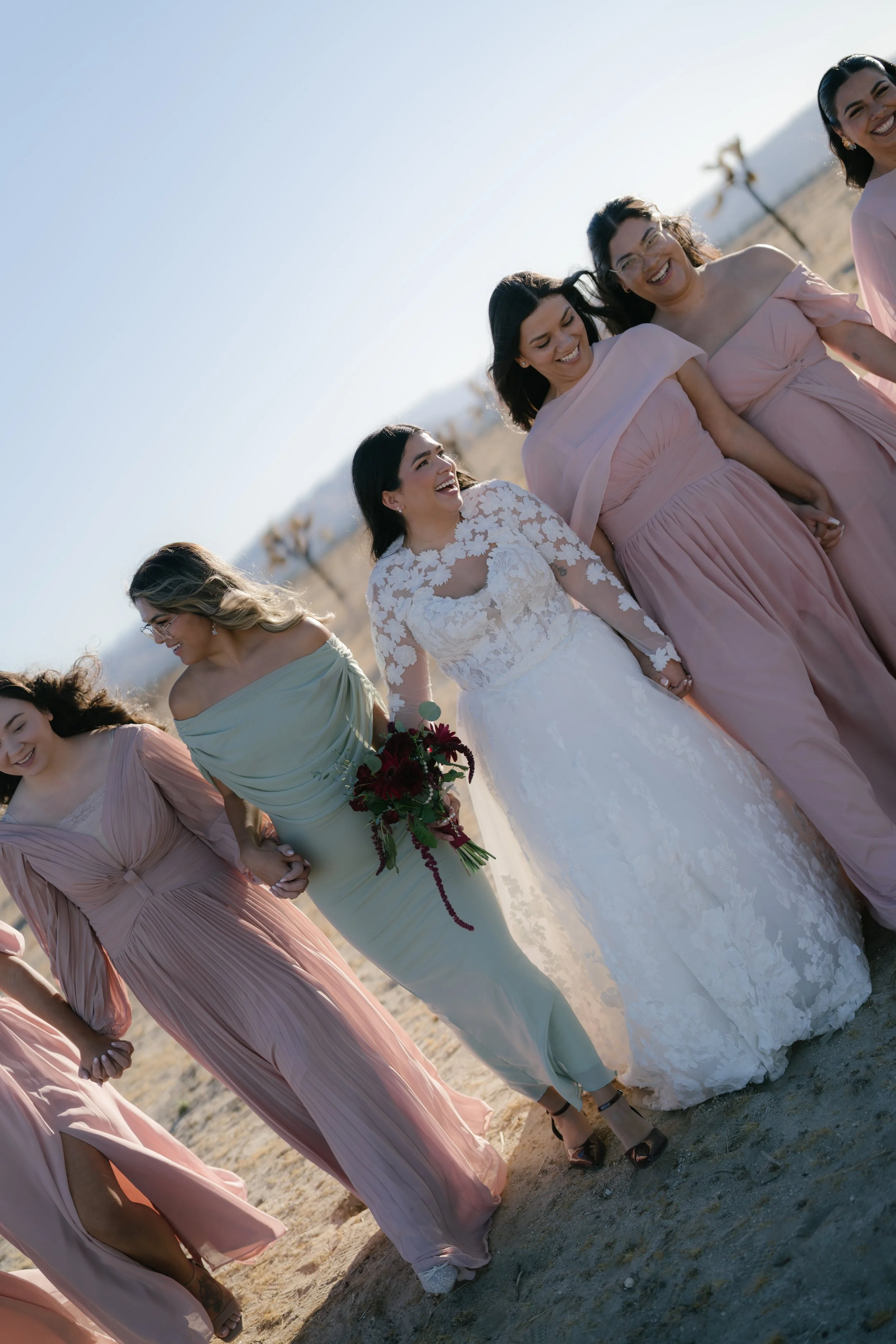 Group of women dressed in pastel-colored formal dresses, walking hand-in-hand on the beach, smiling and enjoying each other's company during sunset.