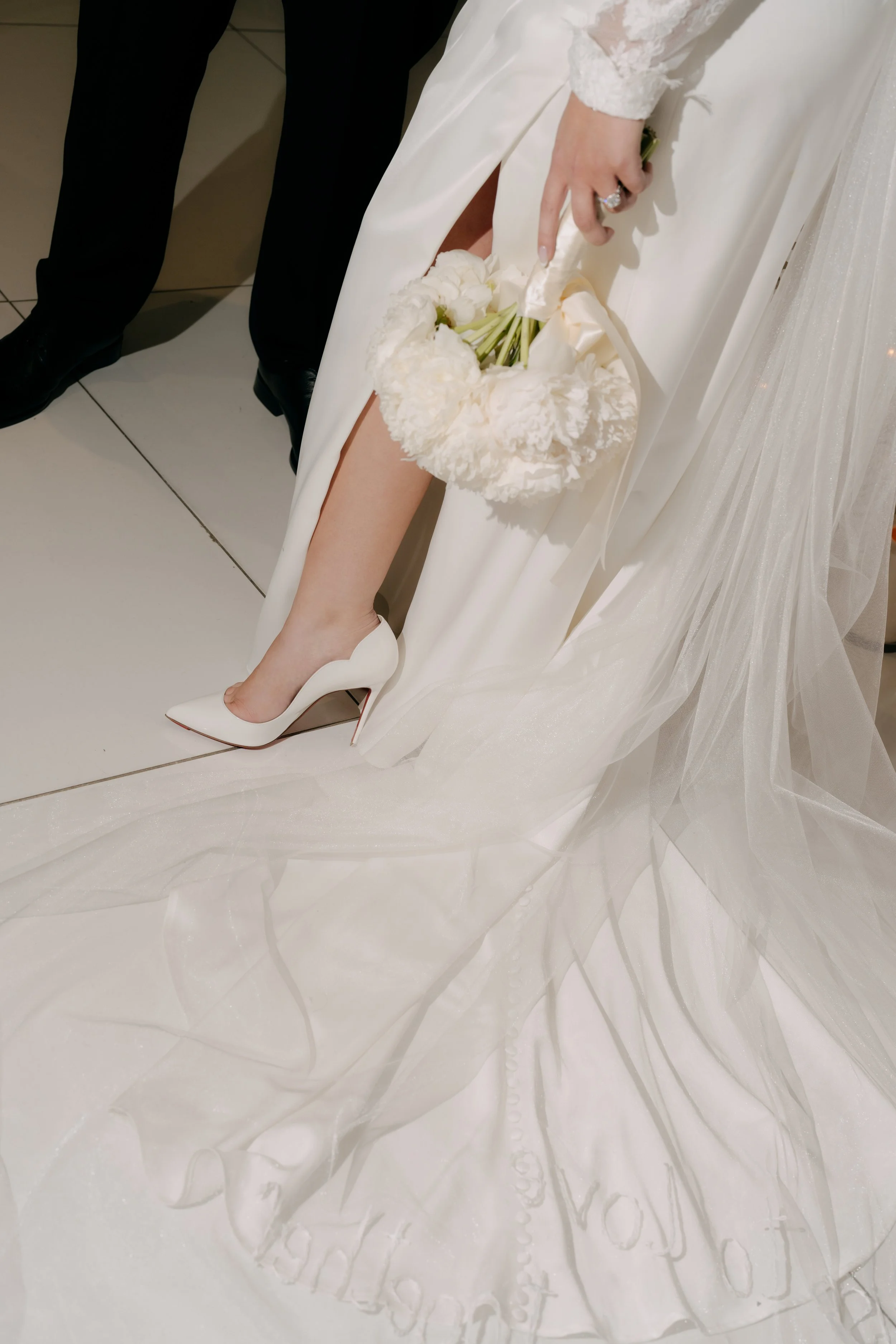 A person wearing a white wedding dress and white high heel shoes, holding a bouquet of white flowers, possibly during a wedding ceremony.