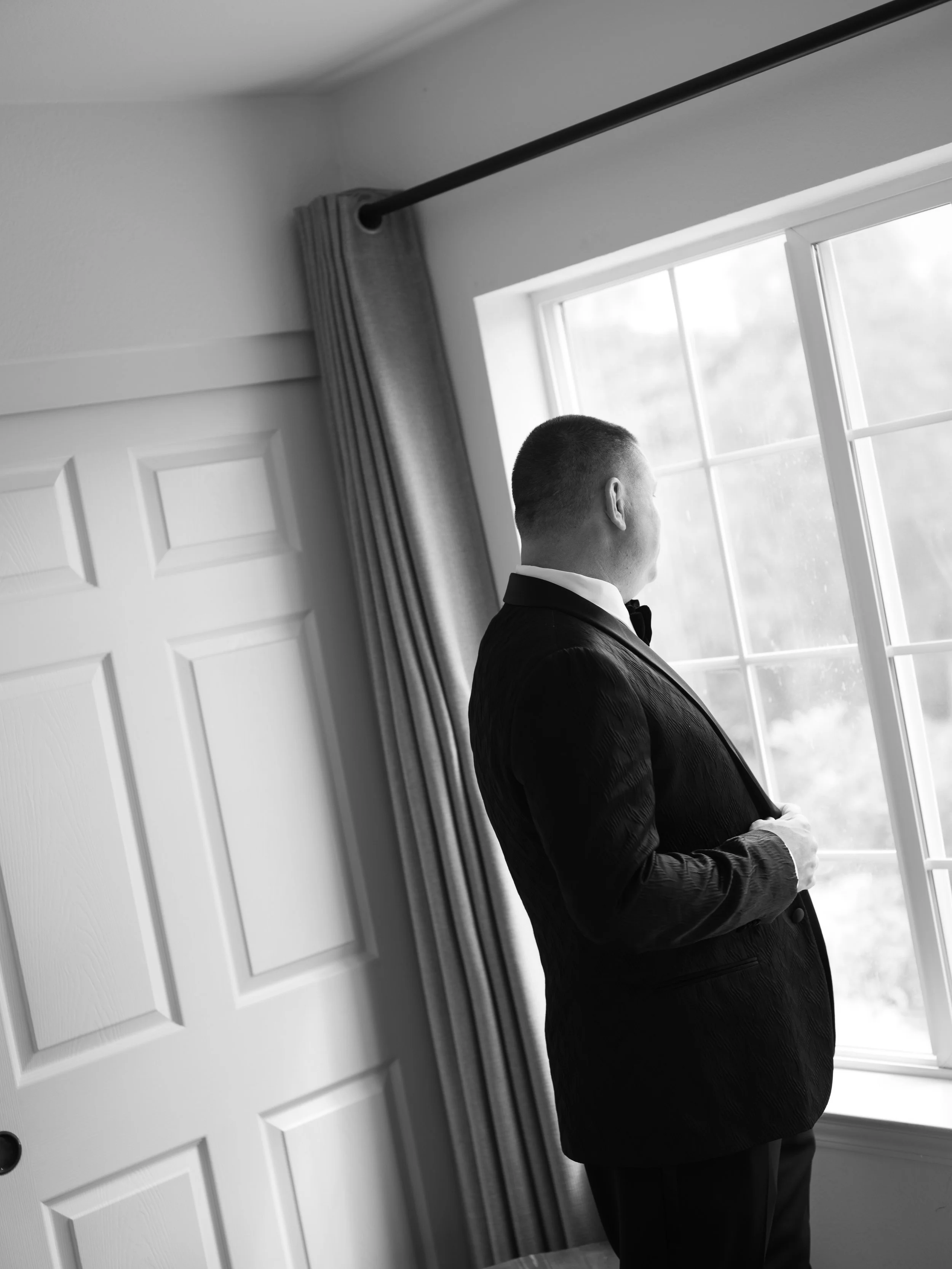 A man in a tuxedo standing by a window, looking outside, with a curtain partially open, in a room with a white door and blinds