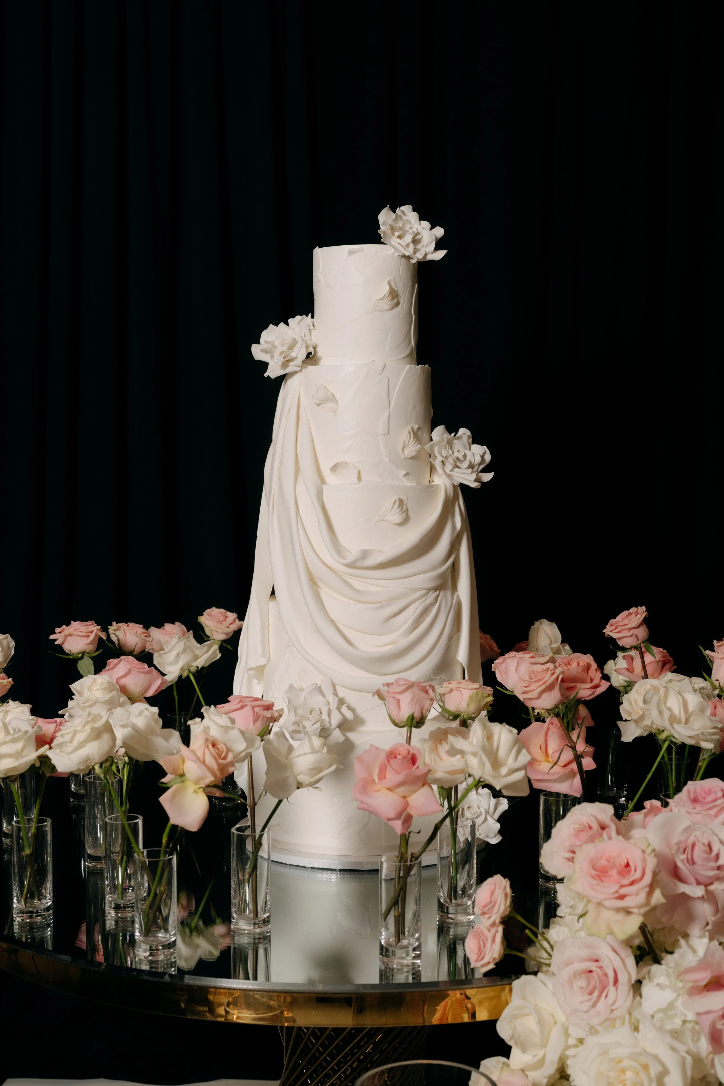 Elegant white three-tiered wedding cake decorated with white flowers and draped icing, surrounded by pink and white roses in clear vases, against a black background.