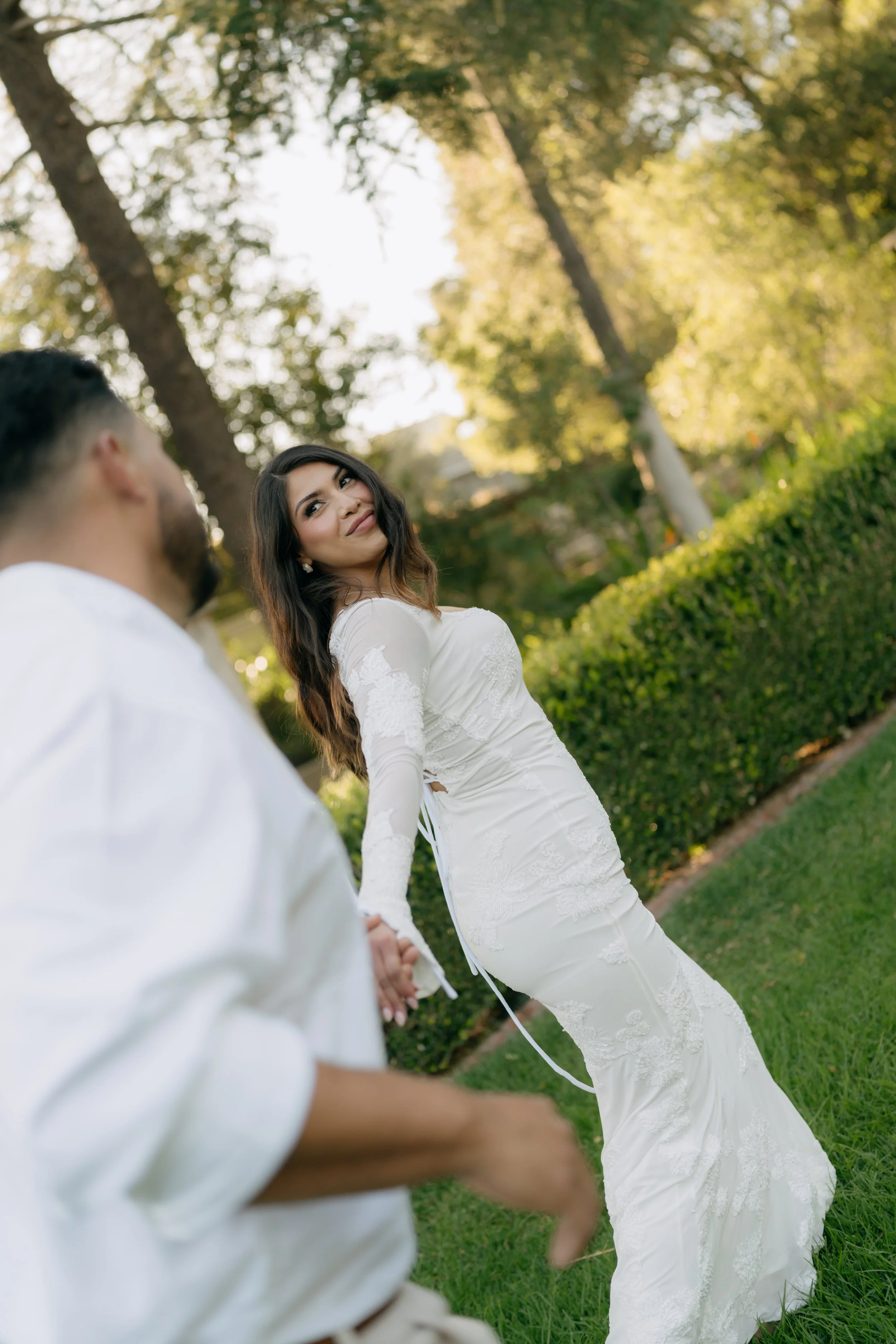 A woman in a white wedding dress holding hands with a man in a white shirt outdoors, with trees and greenery in the background.