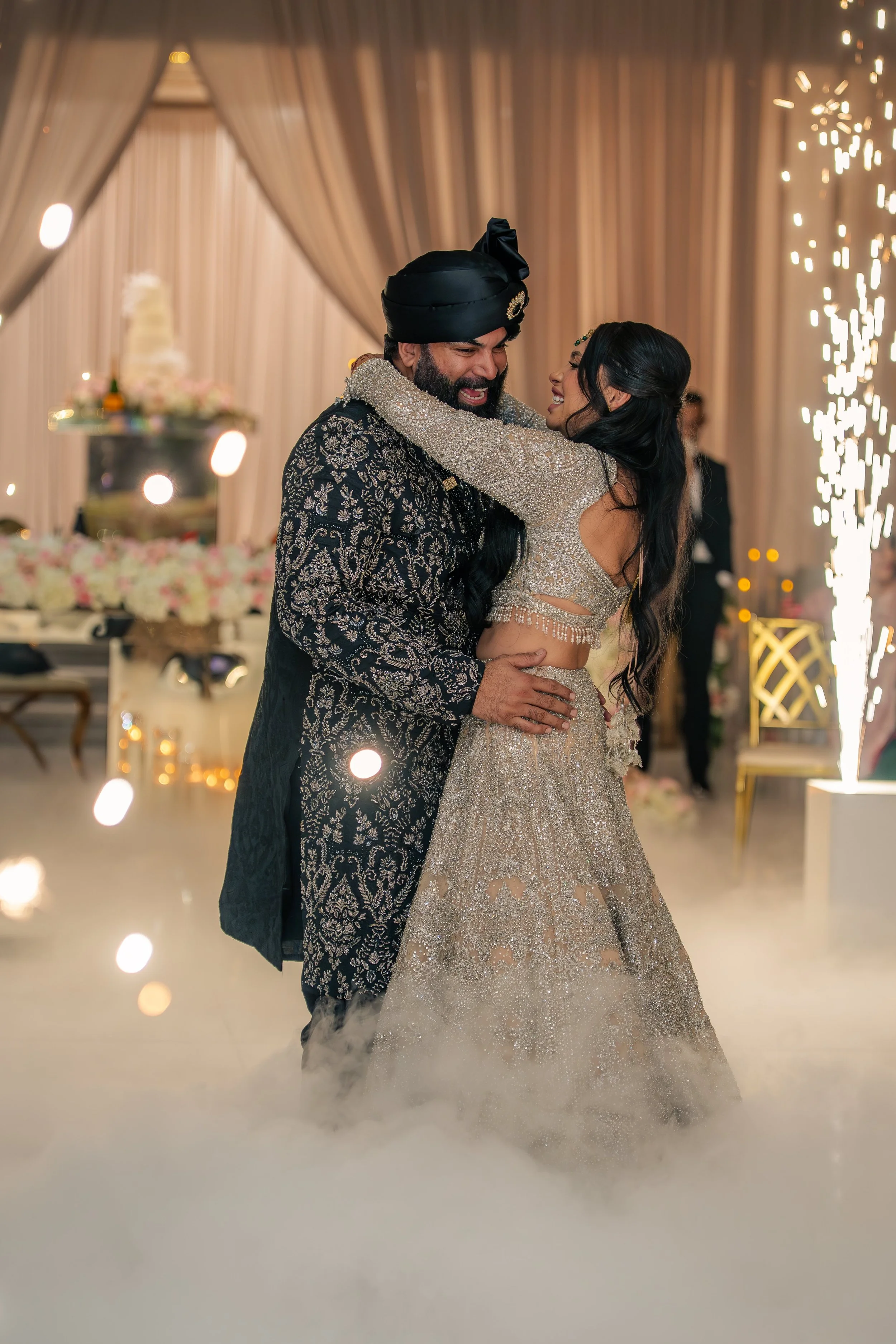 A couple dressed in traditional Indian wedding attire dancing at their wedding reception, with a decorated background and sparklers.