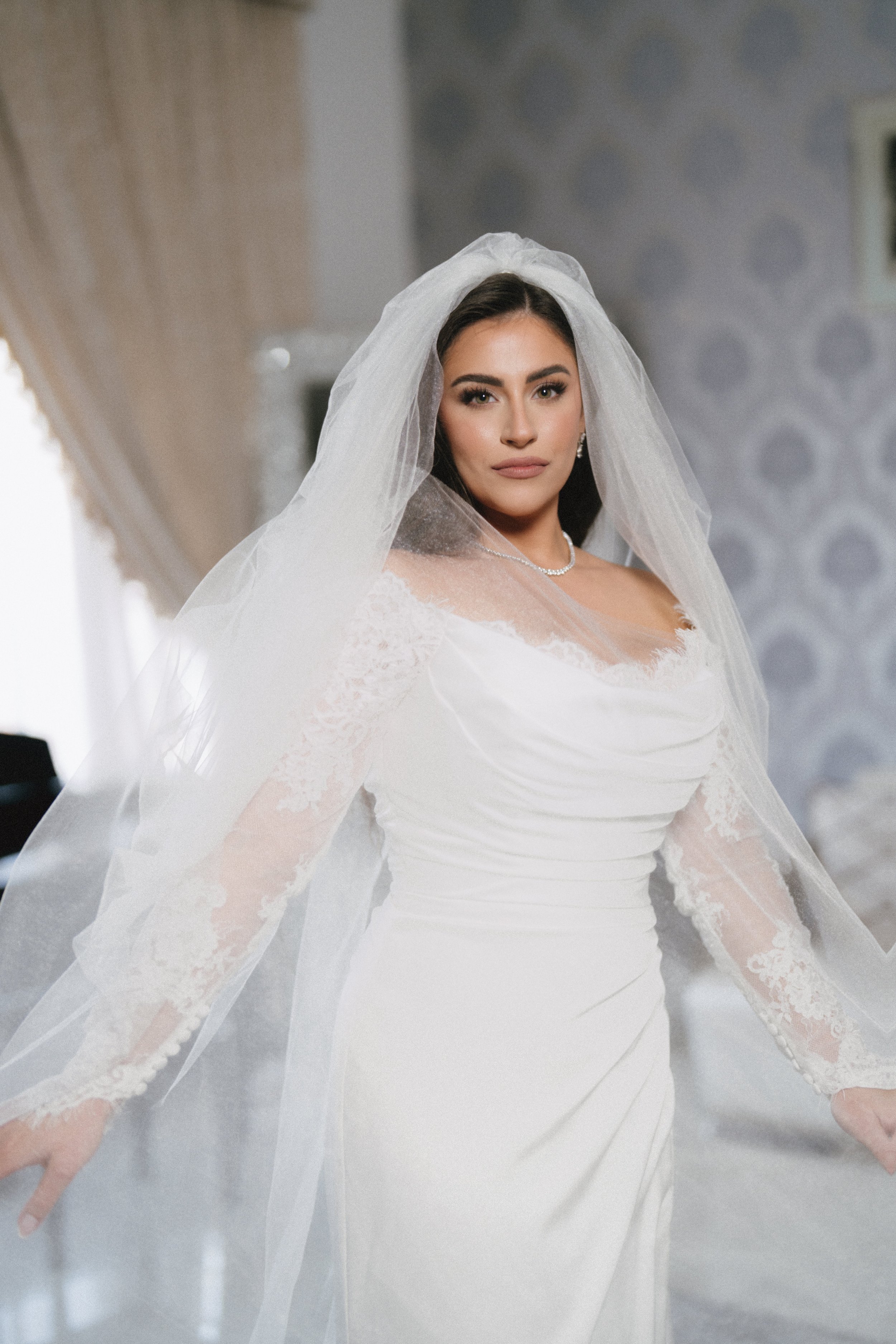 A bride with dark hair, makeup, and jewelry, wearing a white wedding gown with lace sleeves and a long veil, standing indoors with a neutral background.