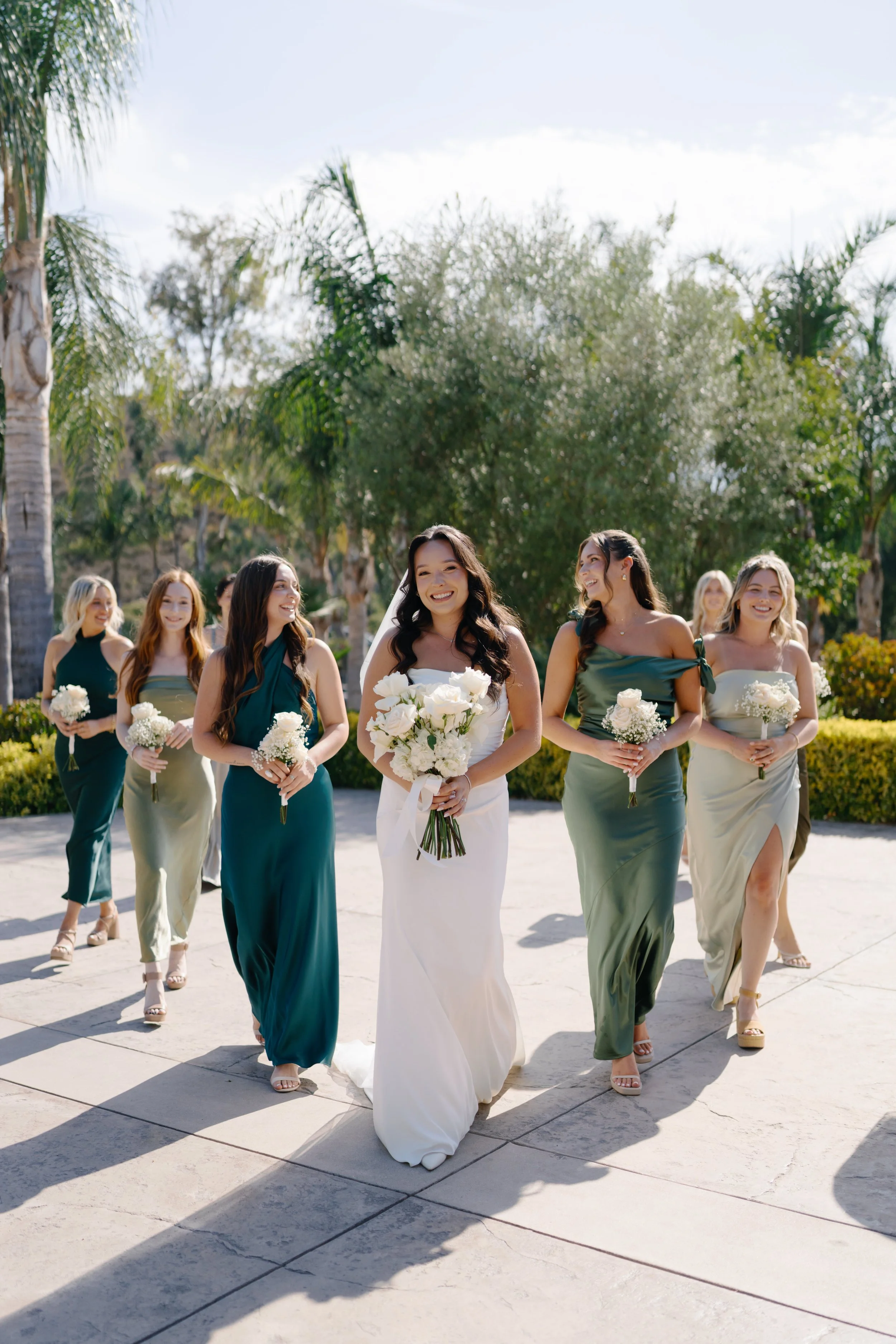 Group of seven women dressed in formal or bridesmaid attire walking outdoors on a sunny day with trees and clear sky in the background.