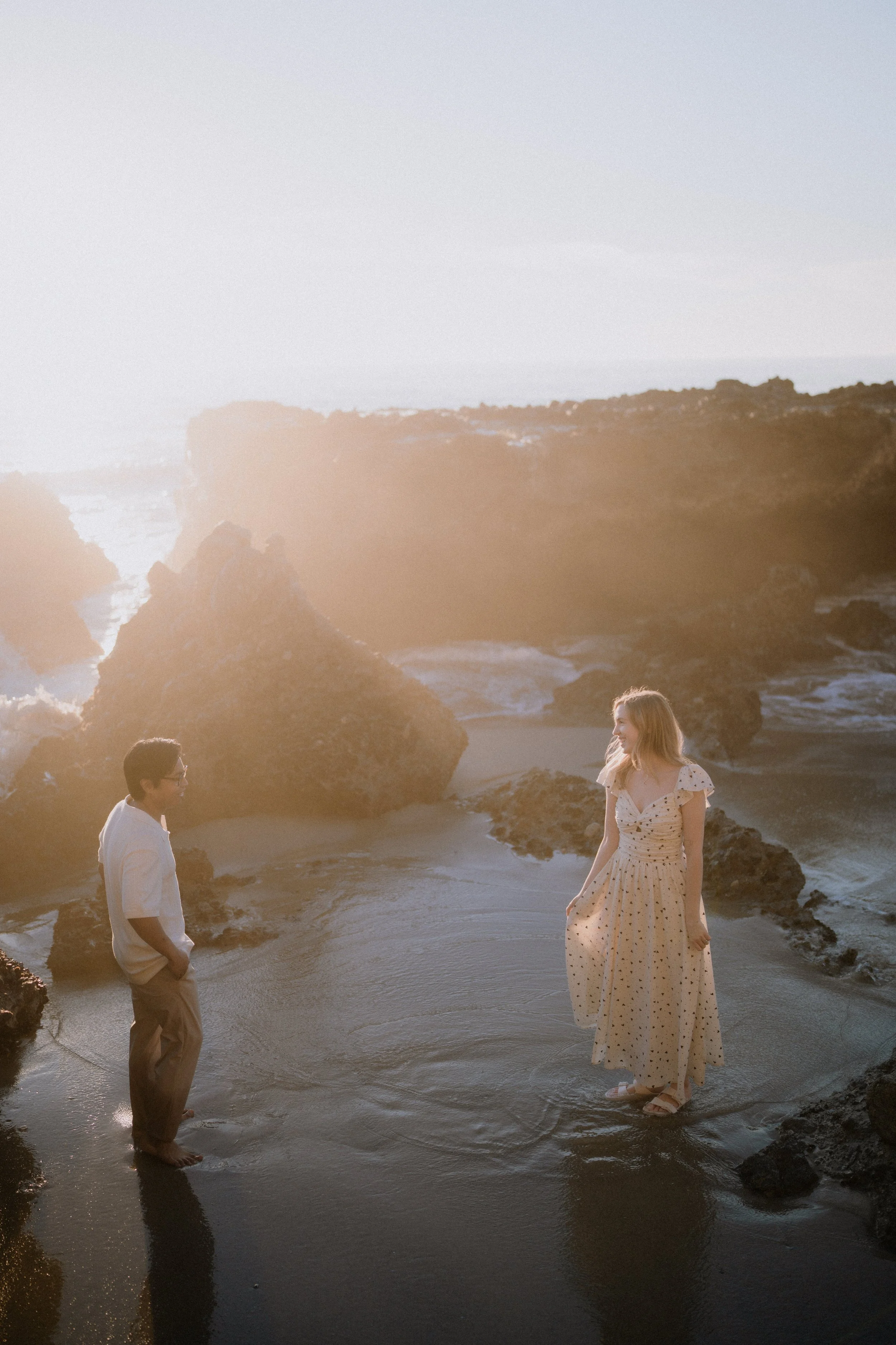 Three people stand on a rocky beach at sunset, with a woman in a long, light-colored dress with polka dots smiling and looking at a man in a white shirt with sleeves rolled up and beige pants who is standing with his hands in his pockets. The ocean, 