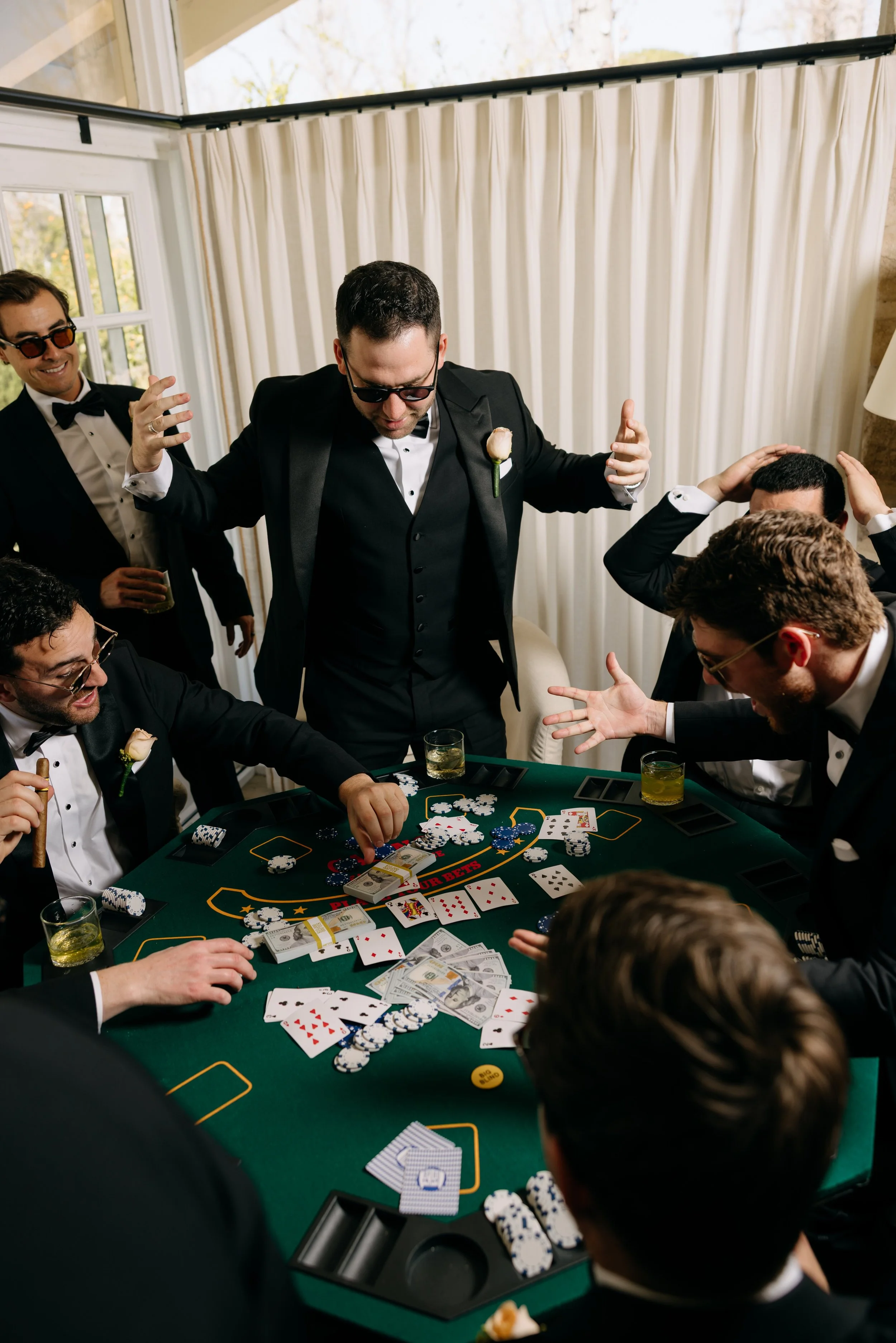 Group of men in tuxedos playing poker at a casino-style table, with drinking glasses and poker chips on the table, in a well-lit room with large windows and white curtains.