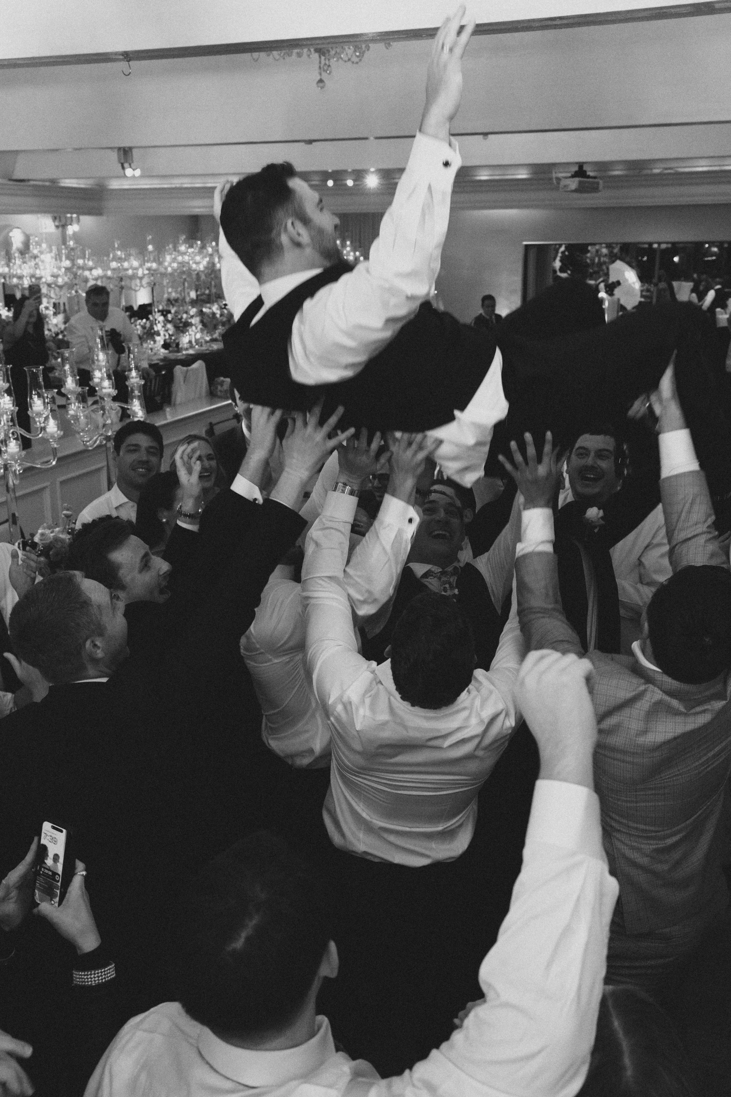 A group of people dressed in formal attire are celebrating at an event, lifting a person above their heads. The person being lifted is wearing a tuxedo and is reaching up with one hand. The background shows a decorated table with candles and a citysc