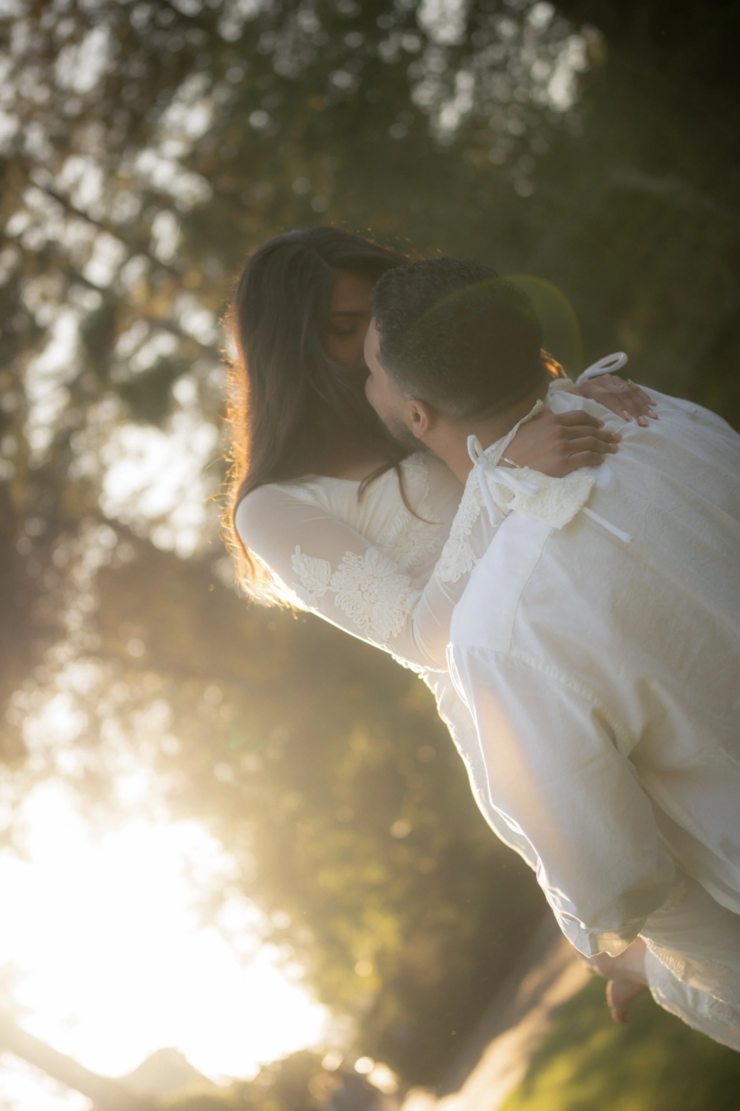 A couple sharing a kiss outdoors during sunset, with the woman wearing a lace white dress and the man in a white shirt, surrounded by trees.