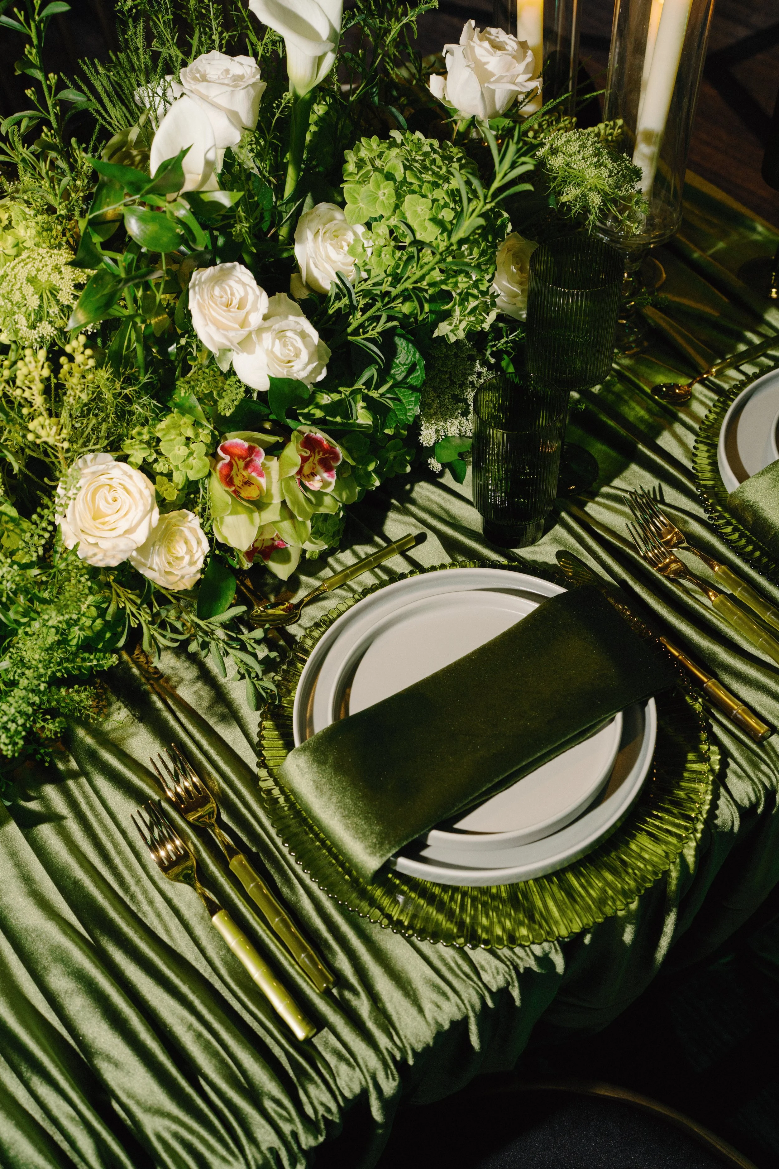 A formal table setting with a green and gold color theme. The table has a green satin tablecloth, gold utensils, and a green glass plate. There is a white plate in the center with a green napkin rolled on top. A floral centerpiece with white roses, g
