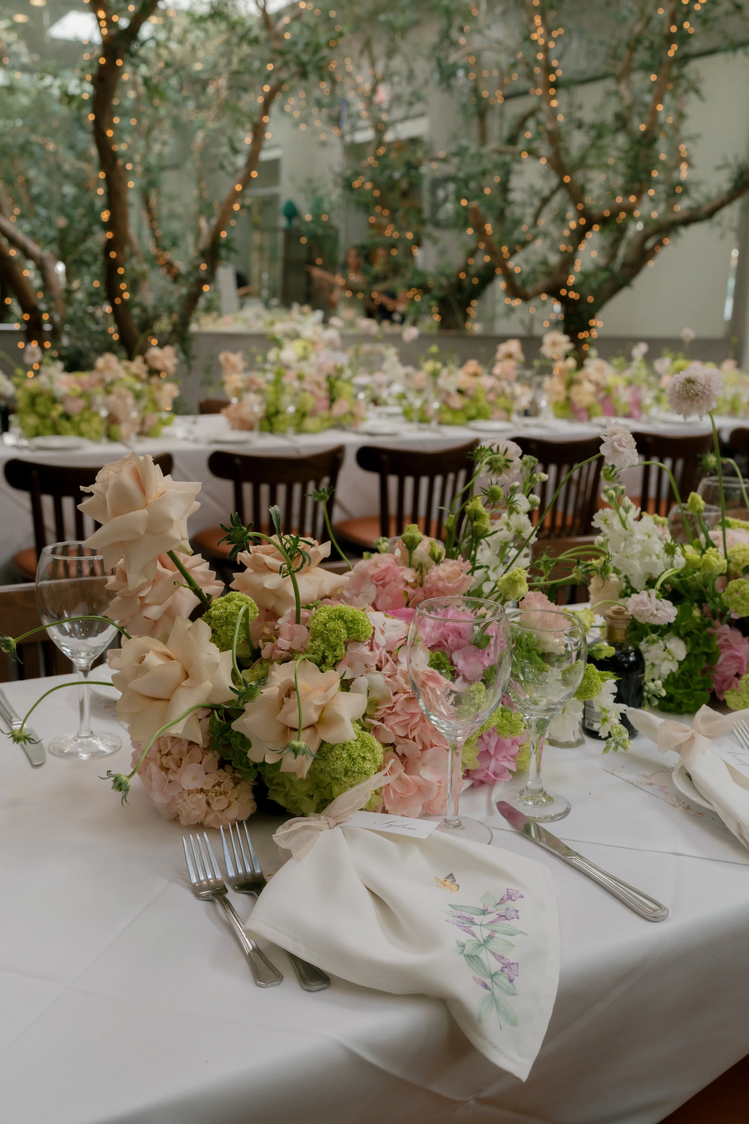 A close-up of a beautifully decorated dining table set for a formal event. The table features a centerpiece of pastel pink, white, and green flowers, with empty wine glasses and silverware arranged neatly. There is a napkin with a floral design, and 