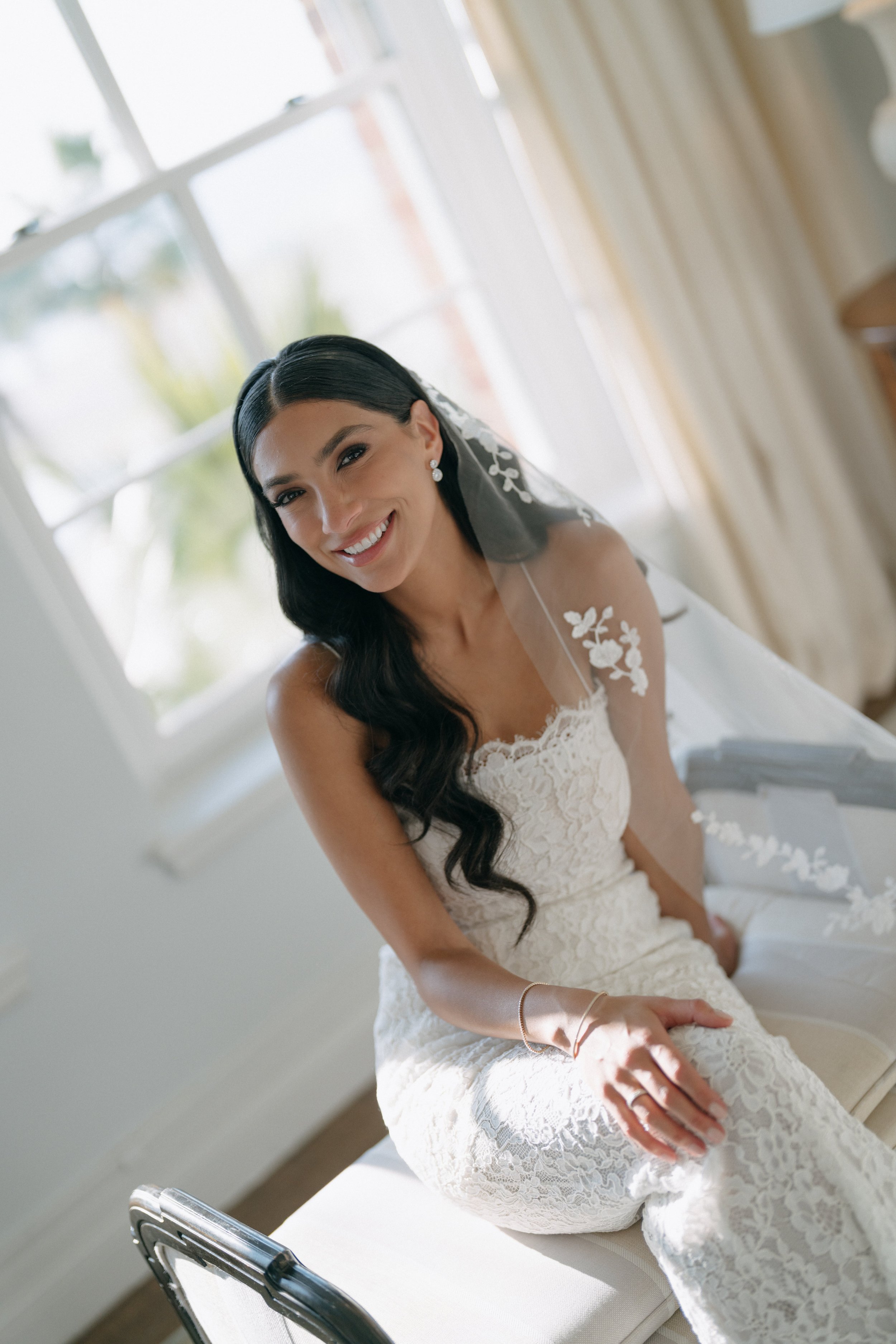 A woman in a white lace wedding dress sitting on a bed near a window, smiling at the camera, with dark hair styled in loose waves.