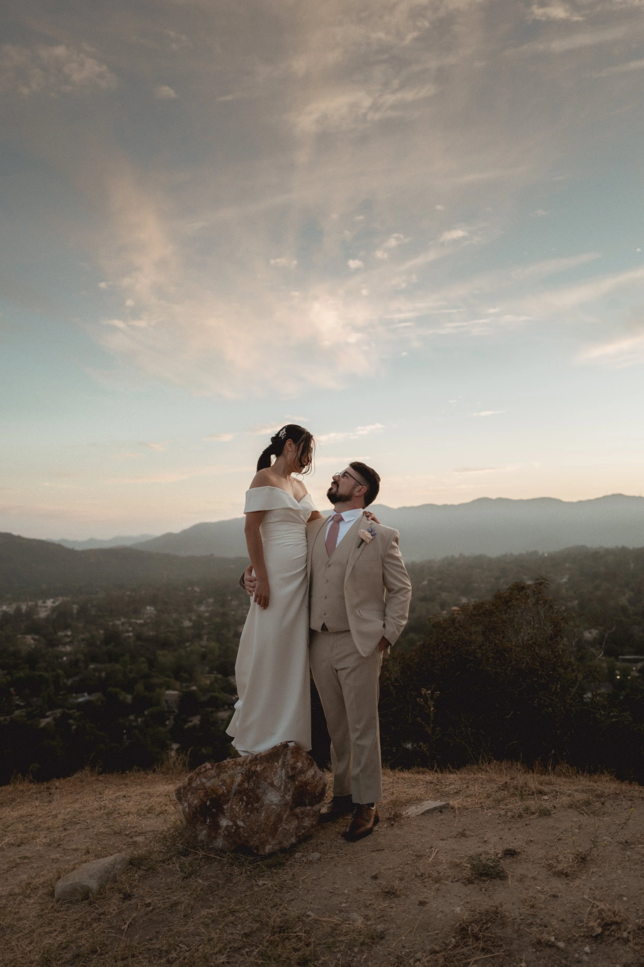 A couple dressed in wedding attire standing outdoors on a hilltop overlooking a valley with mountains in the distance at sunset. The woman is being held up by the man, both looking at each other affectionately.