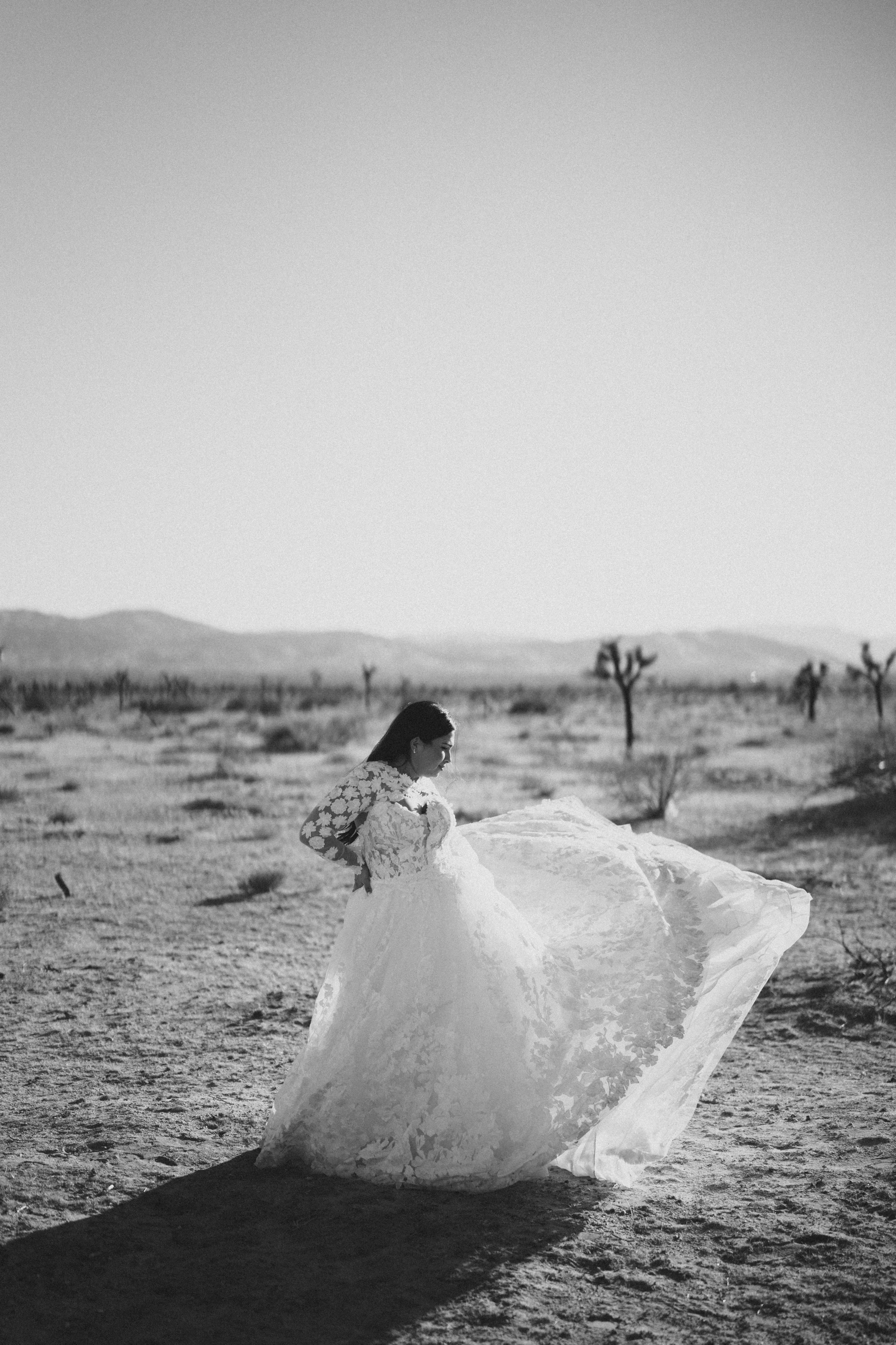 A woman in a wedding dress standing outdoors in a desert landscape with sparse trees and mountains in the background, black and white photo.