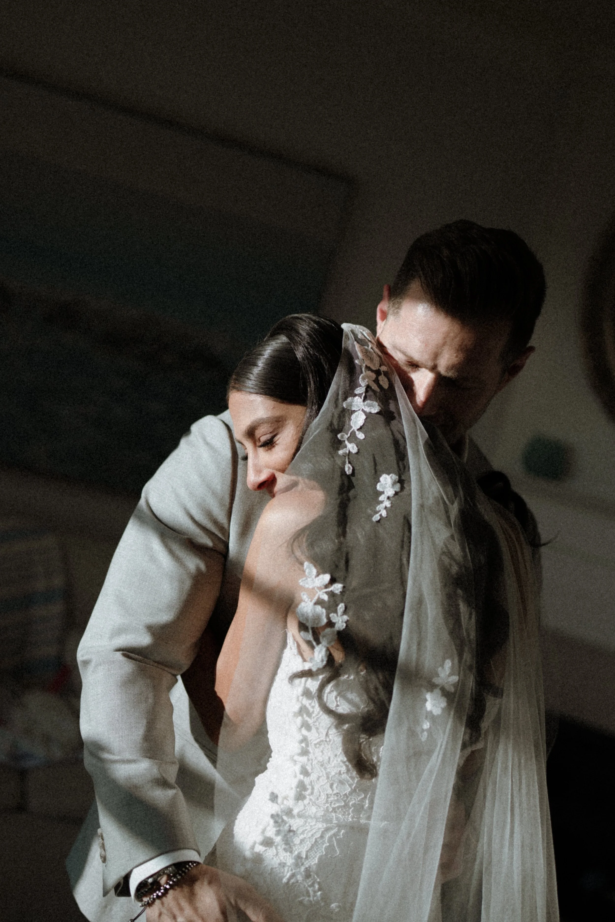 A bride and groom embrace at their wedding, with the bride wearing a white dress with lace detailing and the groom in a gray suit.