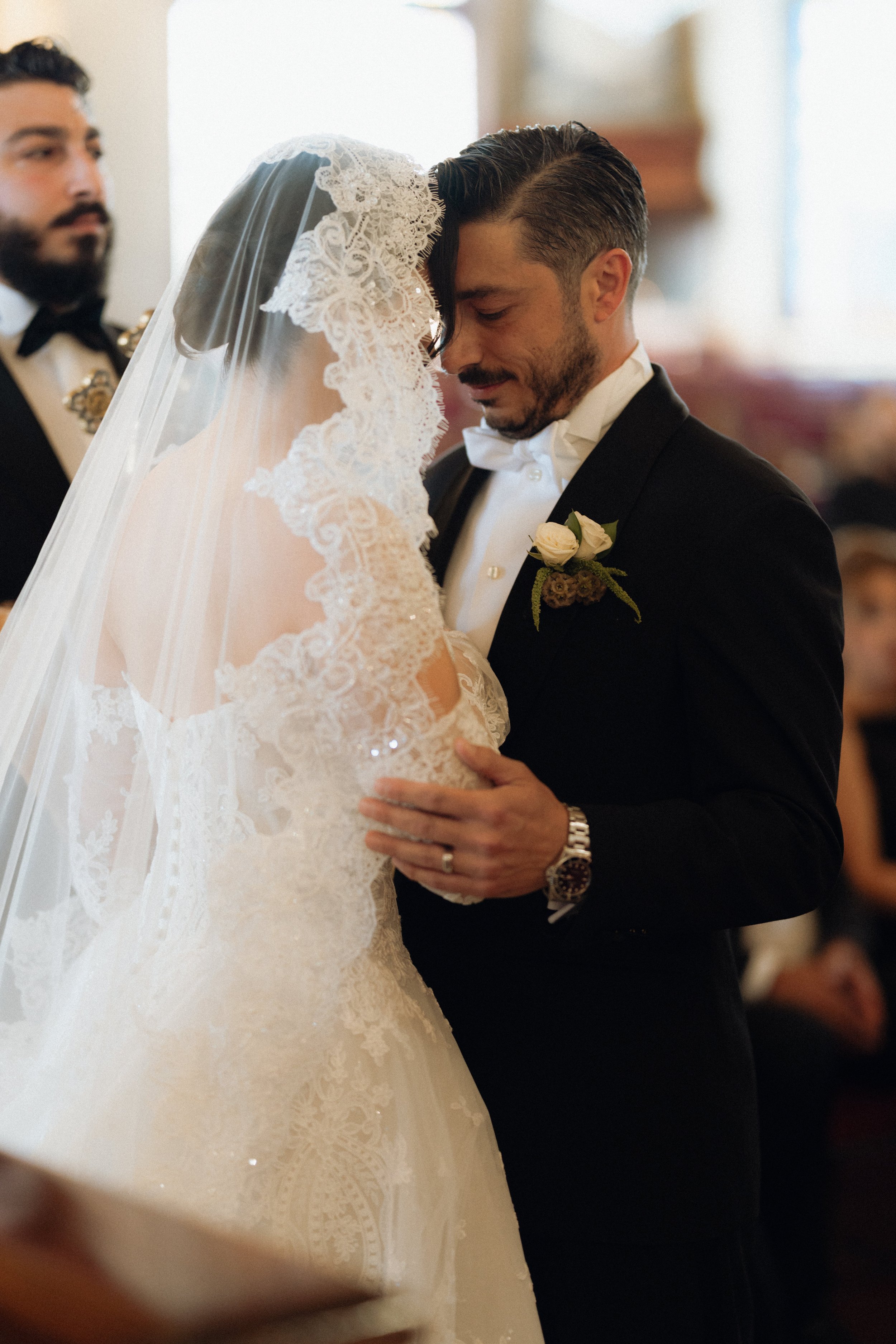 A bride and groom sharing a tender moment during their wedding ceremony, with a third man in the background.