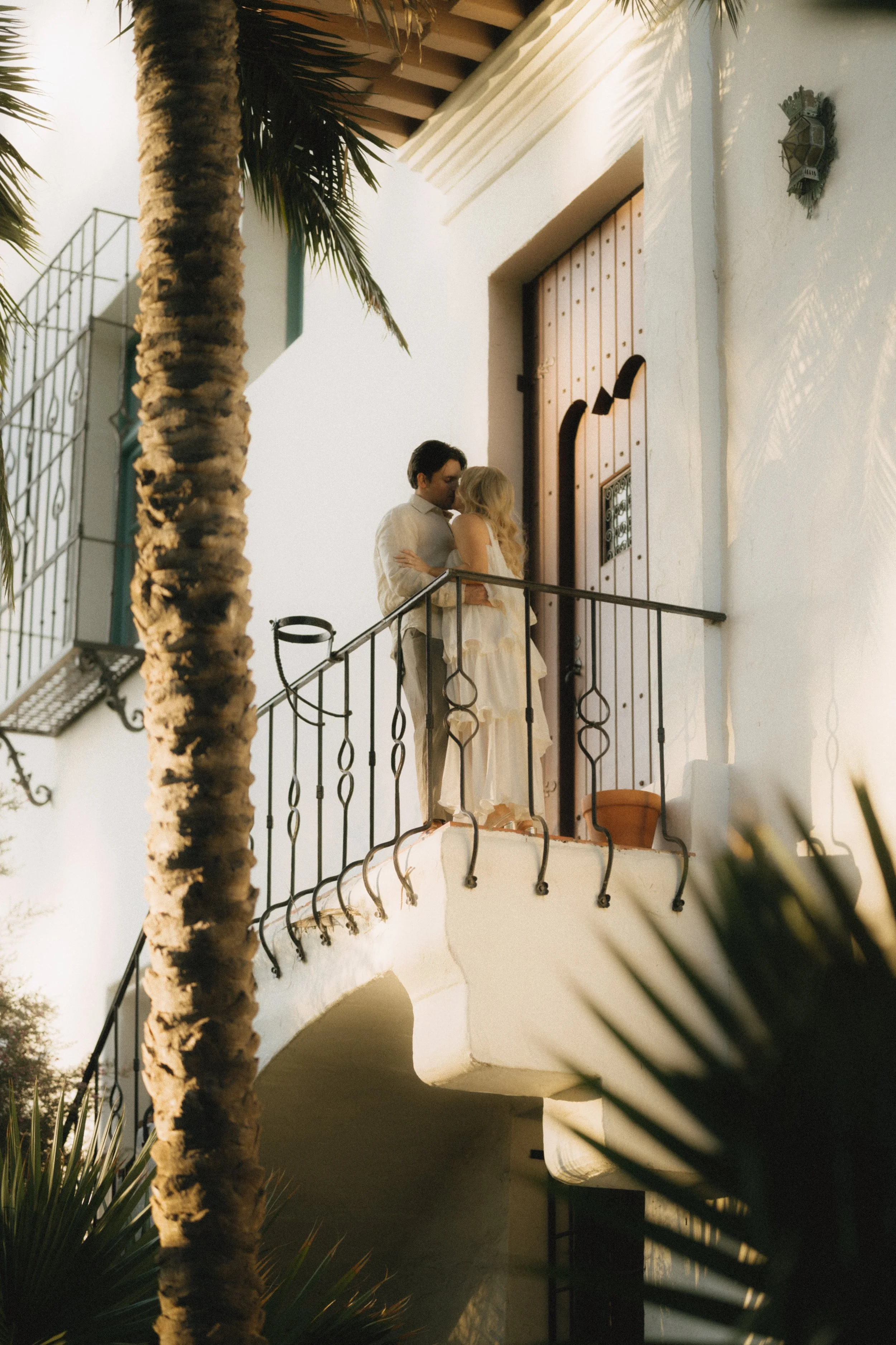 A couple stands on a balcony outside a white Mediterranean-style building during golden hour. The man has dark hair and wears a light-colored shirt; the woman has long blonde hair and wears a light dress. A large palm tree is in the foreground, and s