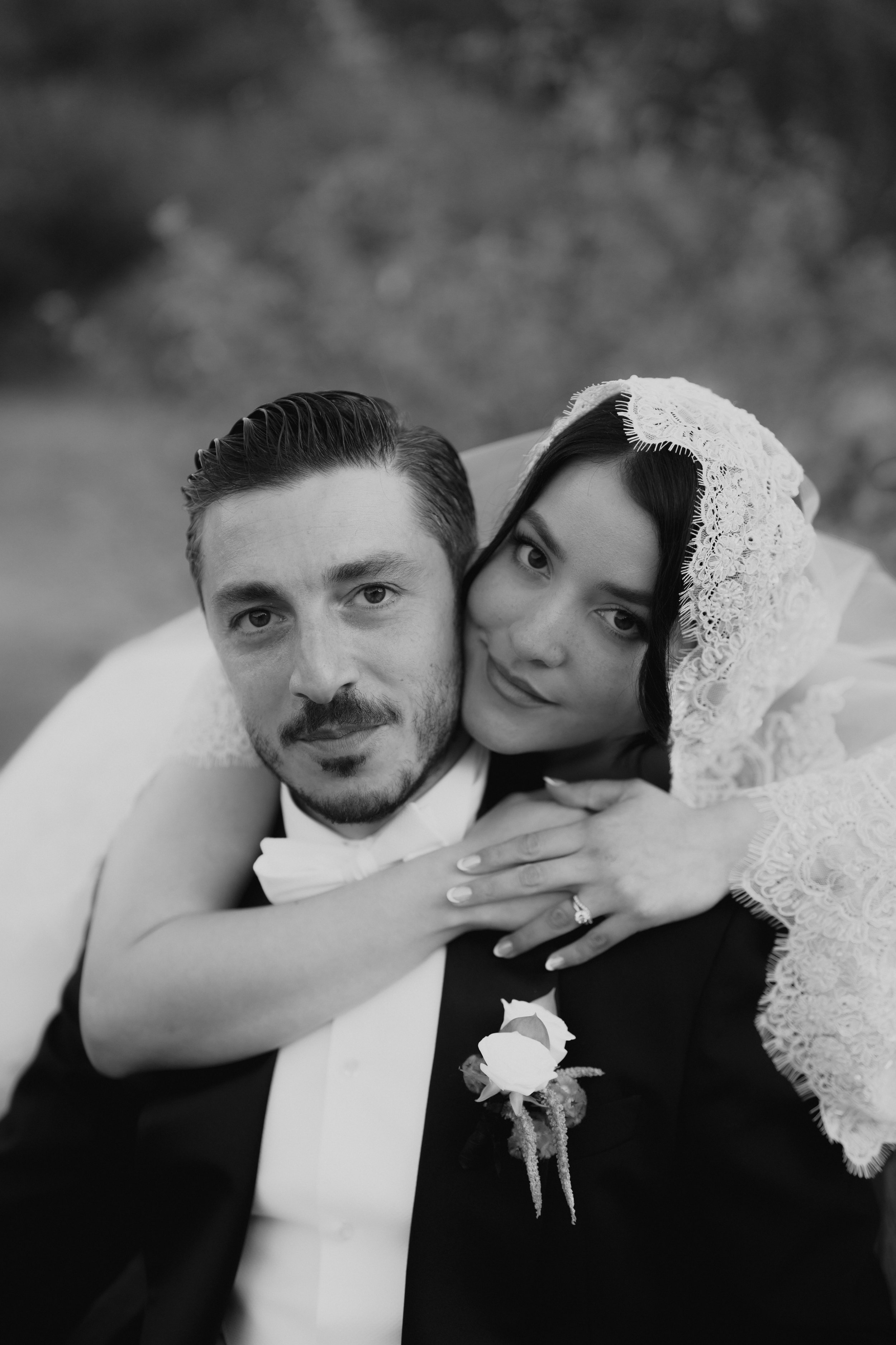 Black and white photo of a newly married couple, with the woman resting her head on the man's shoulder, both smiling softly. The woman is wearing a lace veil, and the man is in a tuxedo with a boutonniere.