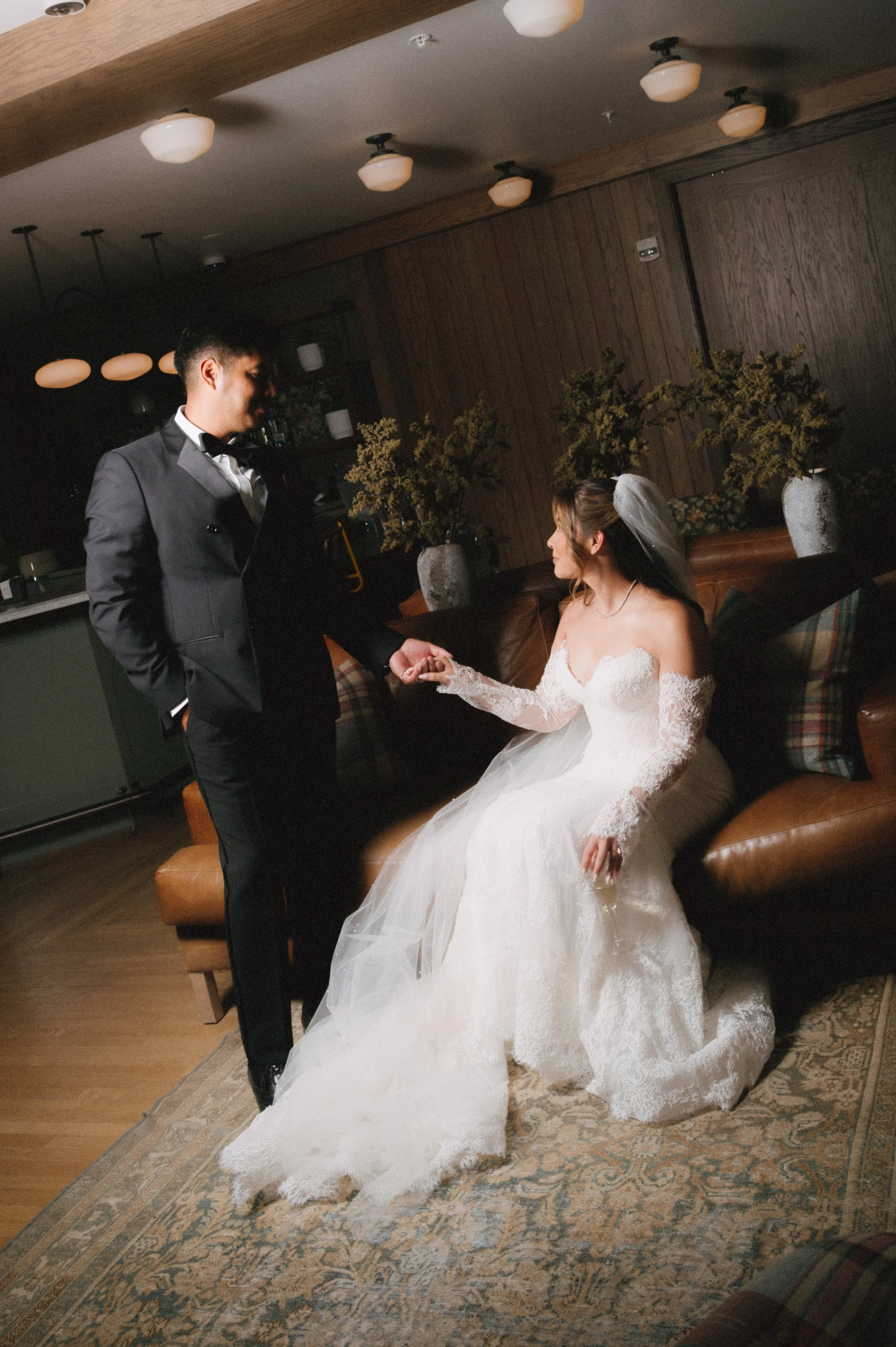 A bride in a white wedding dress sitting on a couch, holding hands with a groom in a black tuxedo standing next to her, indoors with wooden walls and decorative plants.