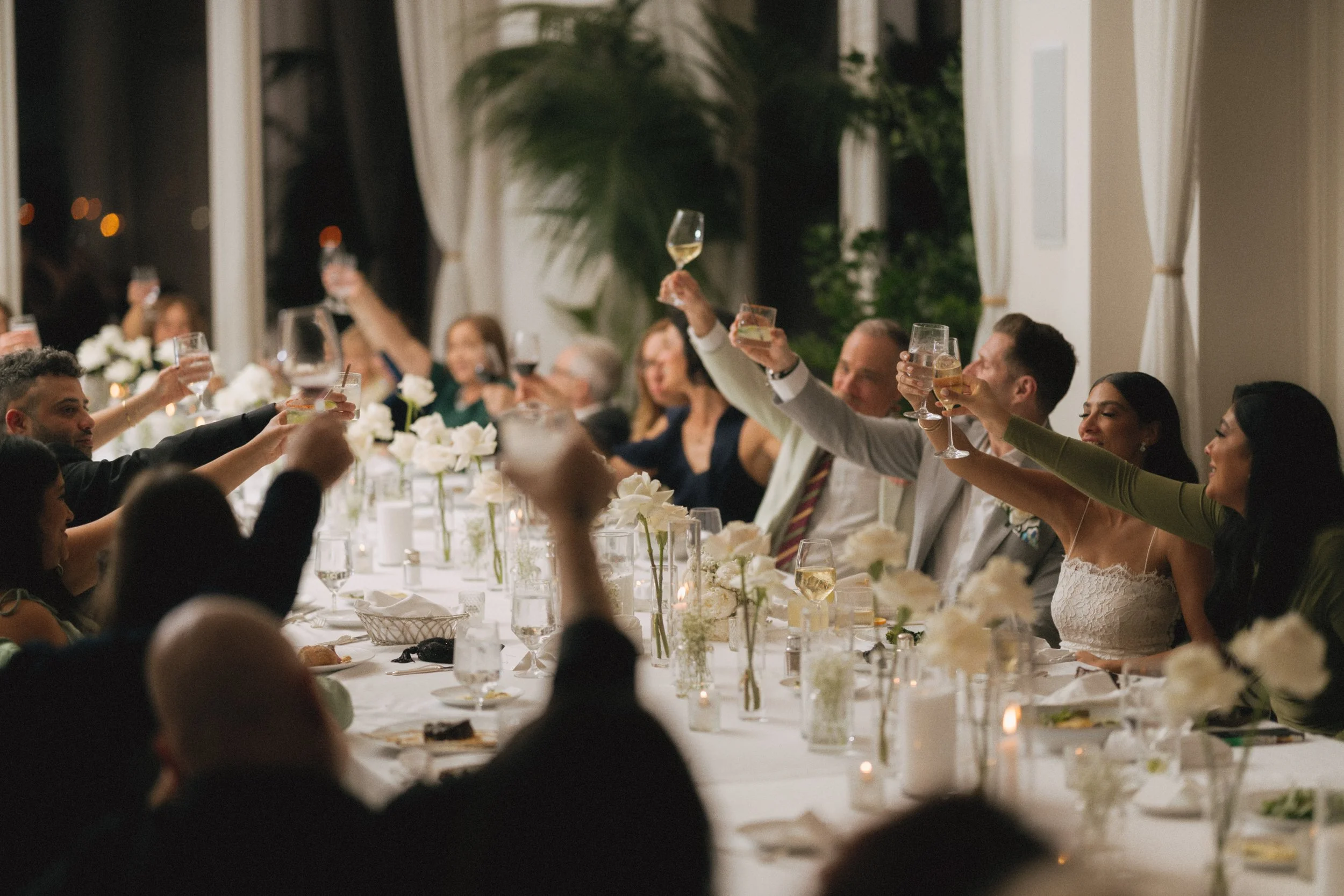 People at a formal dinner raising glasses for a toast, with a long table set with white flowers, candles, and glassware, in a well-lit indoor setting.