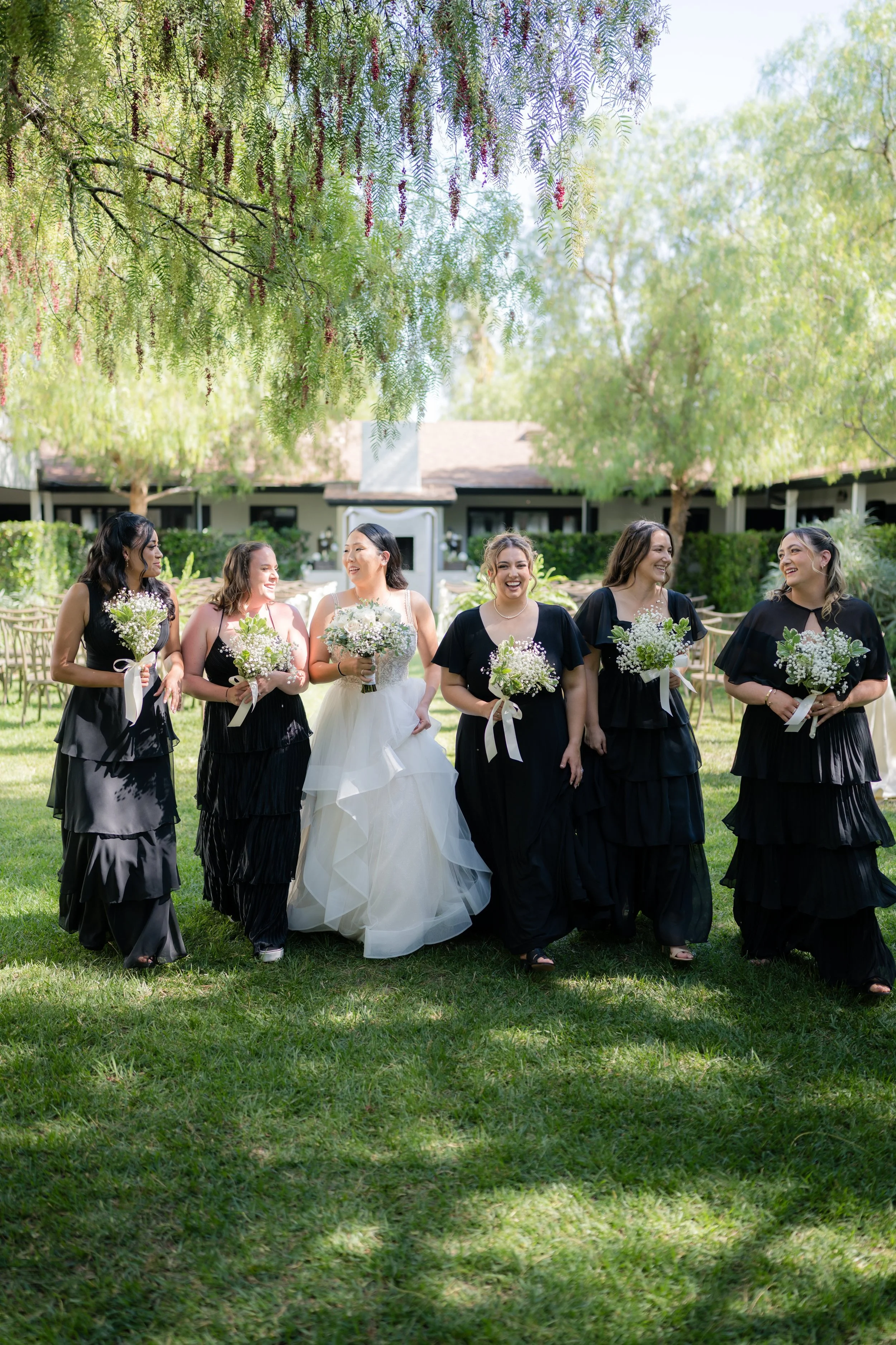 A bride and five women dressed in black walking together outdoors on a sunny day, holding bouquets of flowers, with trees and a house in the background.